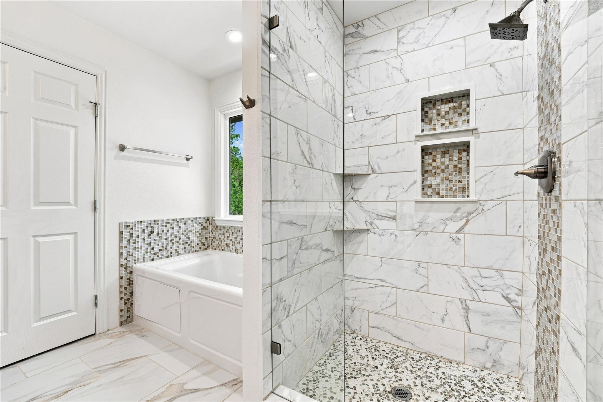 Bathroom with marble-look tile, a glass shower, pebble floor, and white bathtub.