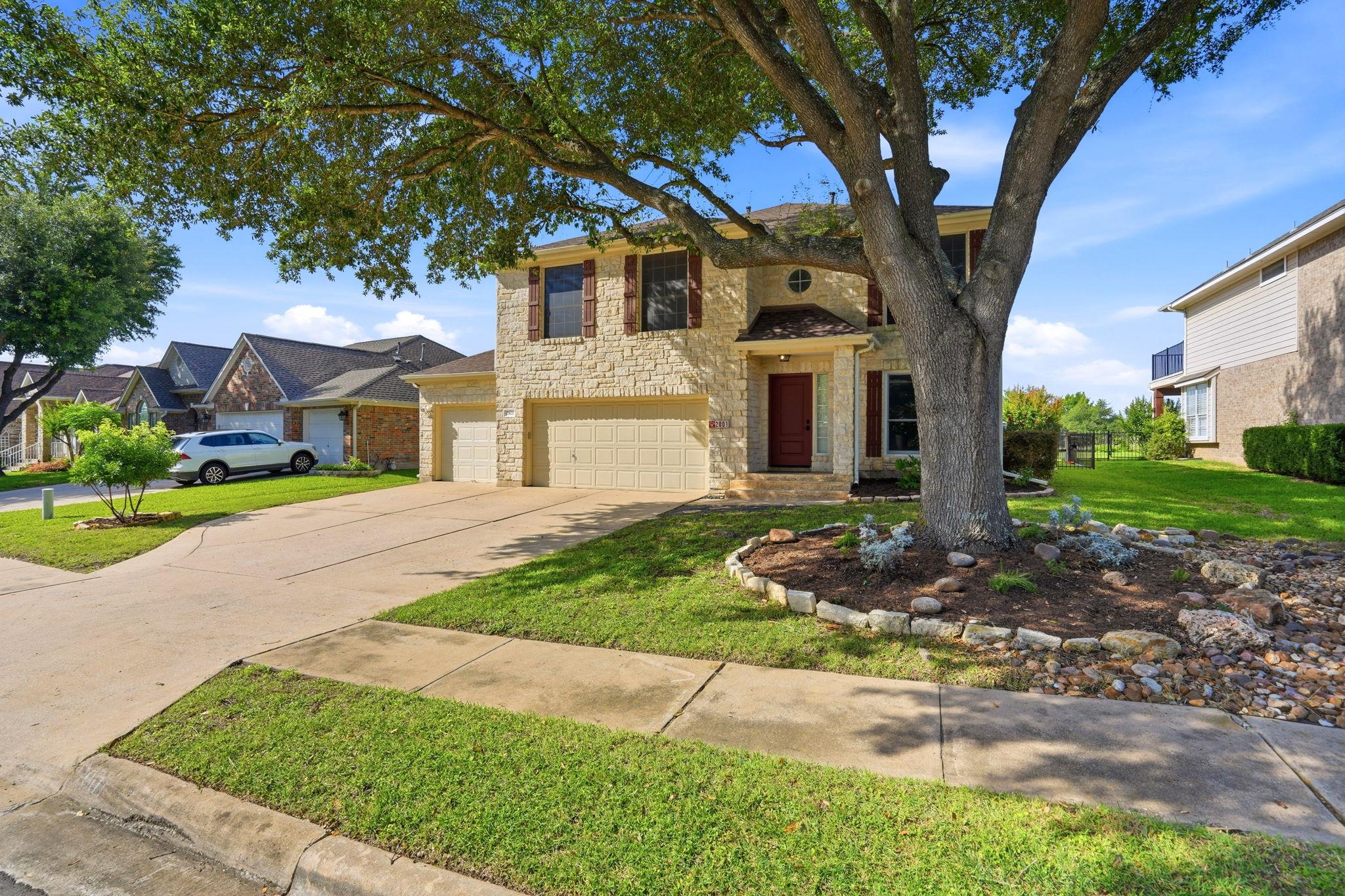 Two-story stone-front home with attached garage, broad driveway, and a large oak tree in the front yard.