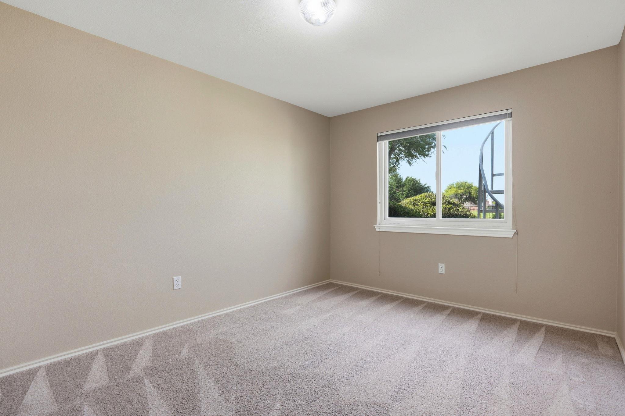 Empty bedroom with beige walls, carpet, and a window overlooking greenery.