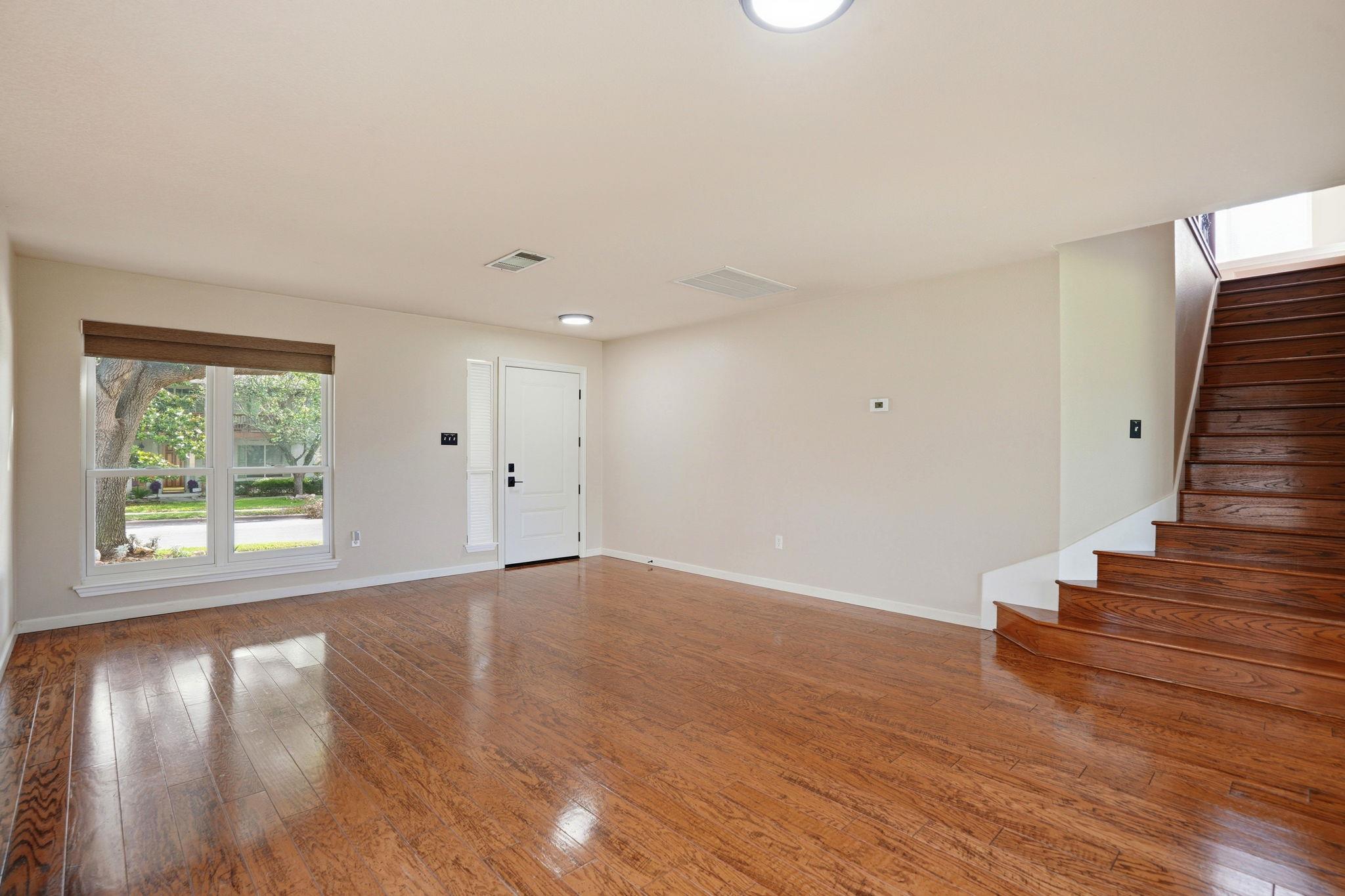 Empty living room with glossy wood floors, a large window, and a dark wood staircase.