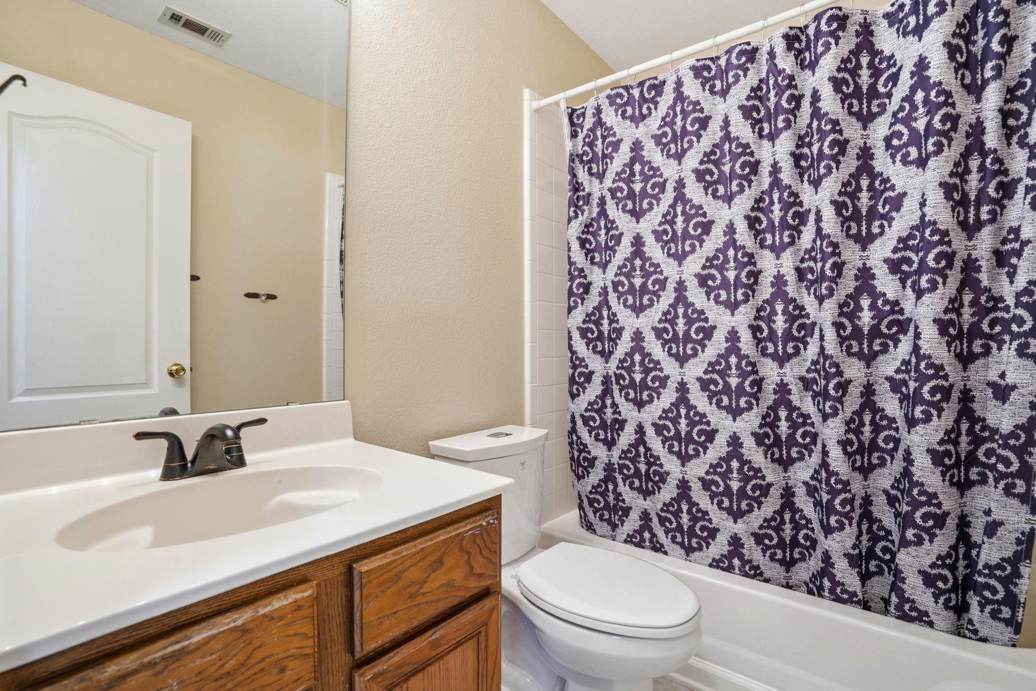 Bathroom with a wood-front vanity, white fixtures, and purple patterned shower curtain.