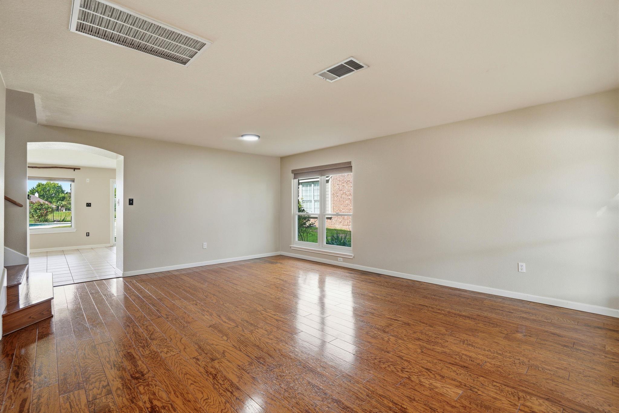 Empty living room with glossy wood floors, a large window, and an arched opening to a tiled area.