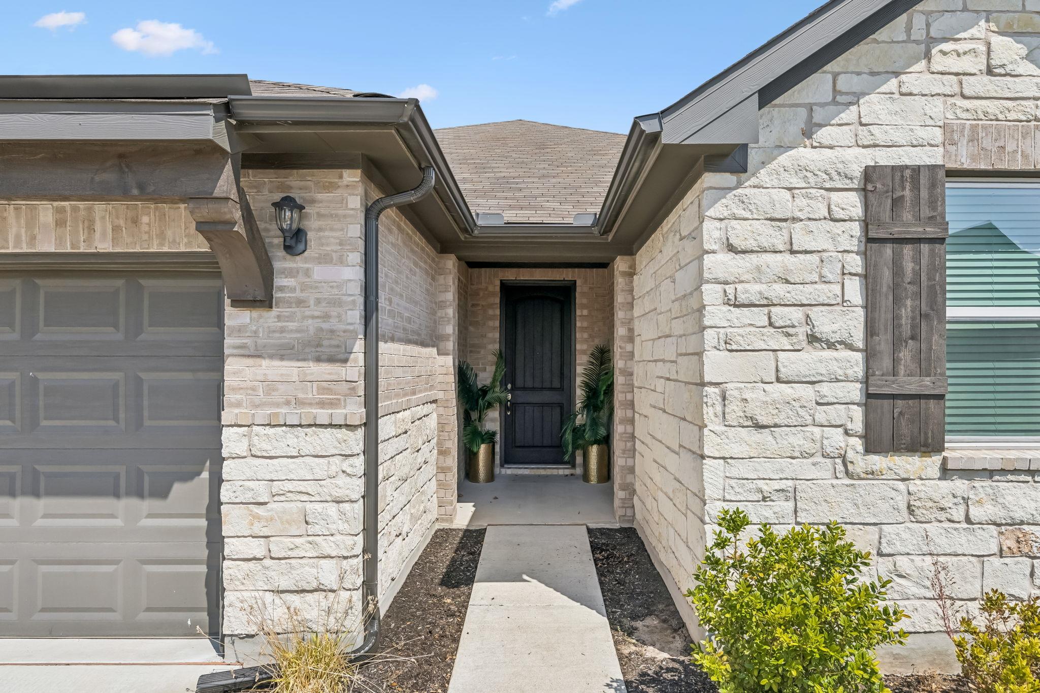Stone-and-brick house entry with a recessed dark door, potted plants, and an attached garage.