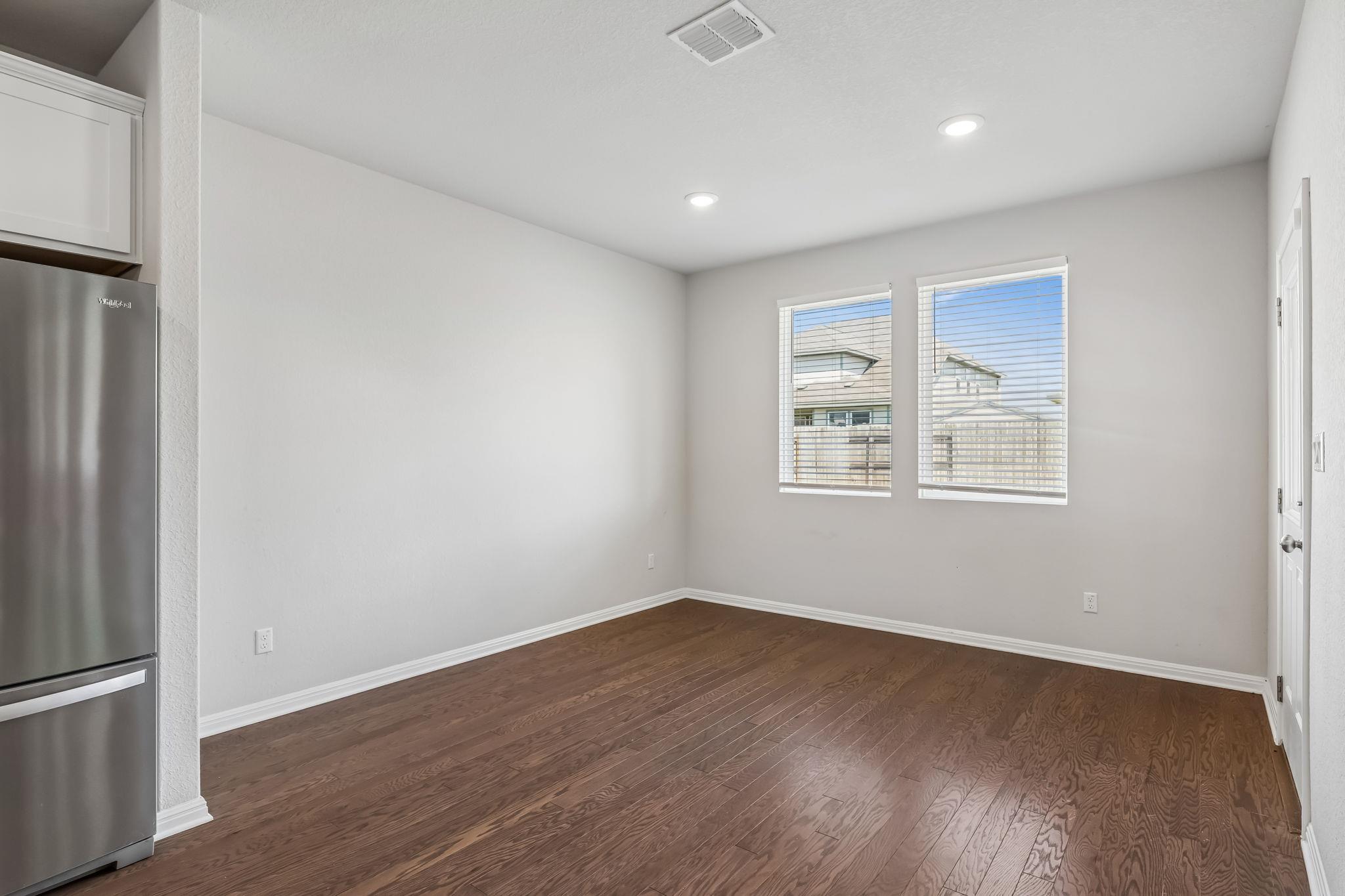 Empty dining area with dark wood floors, two windows, and a stainless steel refrigerator.