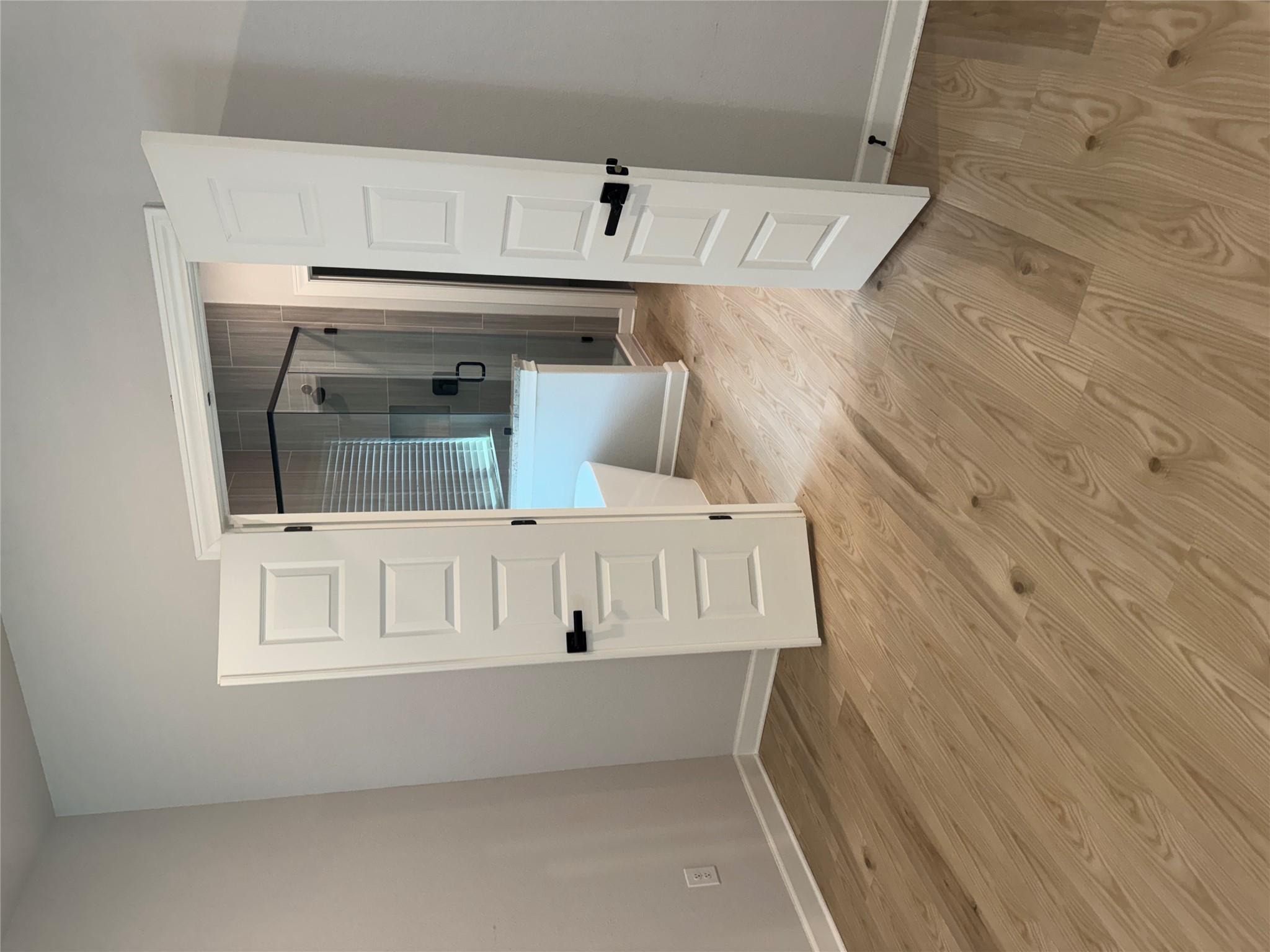 Open double doors reveal a tiled glass shower and partial bathtub beyond a light wood floor.