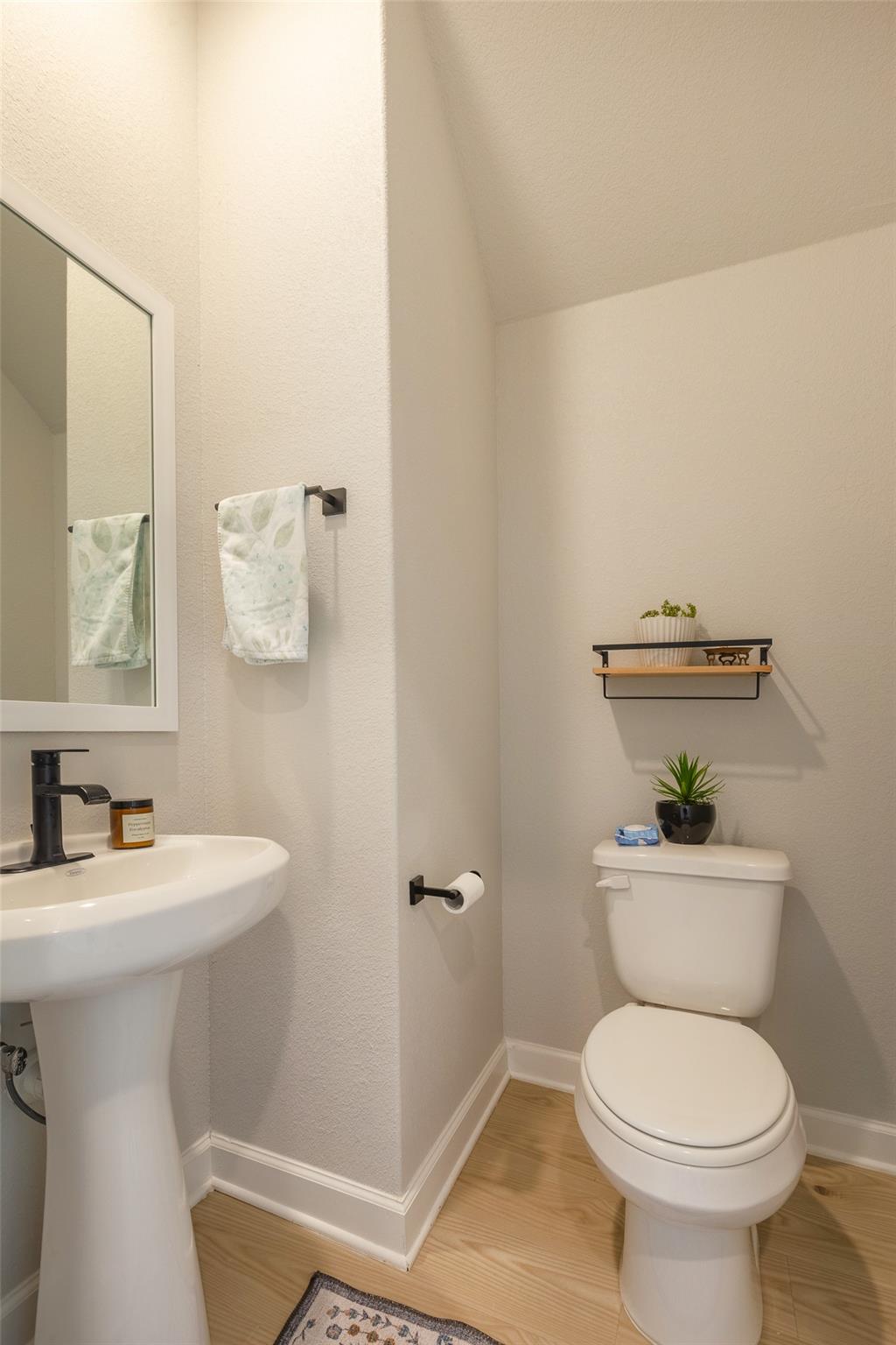 Bathroom with a pedestal sink, framed mirror, and toilet beneath a narrow shelf.