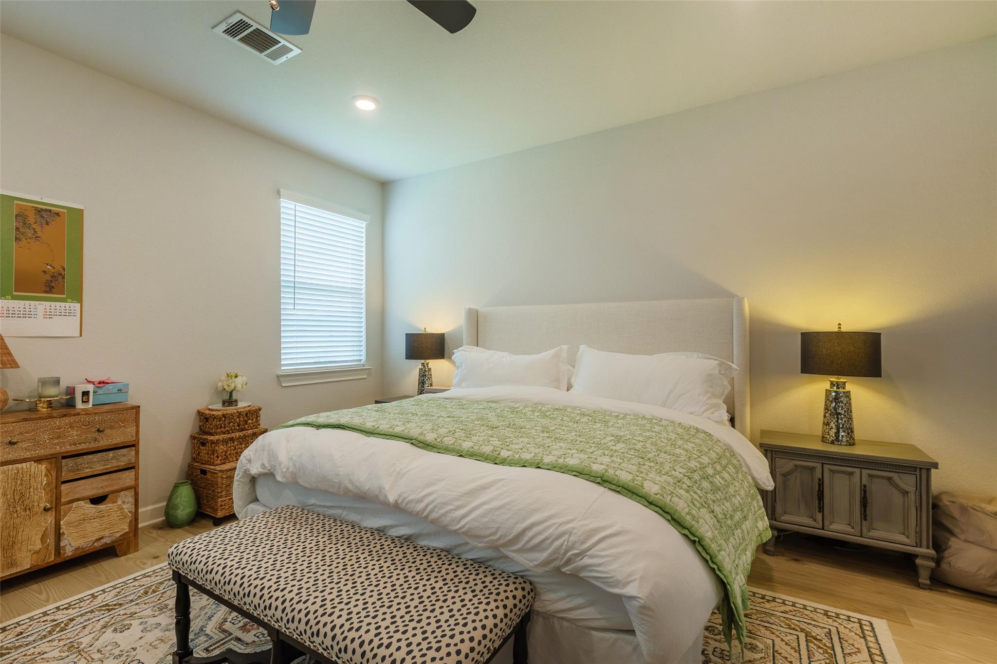 Bedroom with a large white bed, green quilt, patterned bench, and matching bedside lamps.