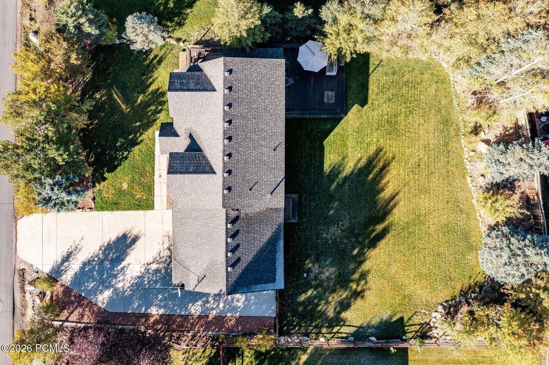 Aerial view of a gray-roofed home, spacious lawn, paved driveway, and deck among autumn trees.