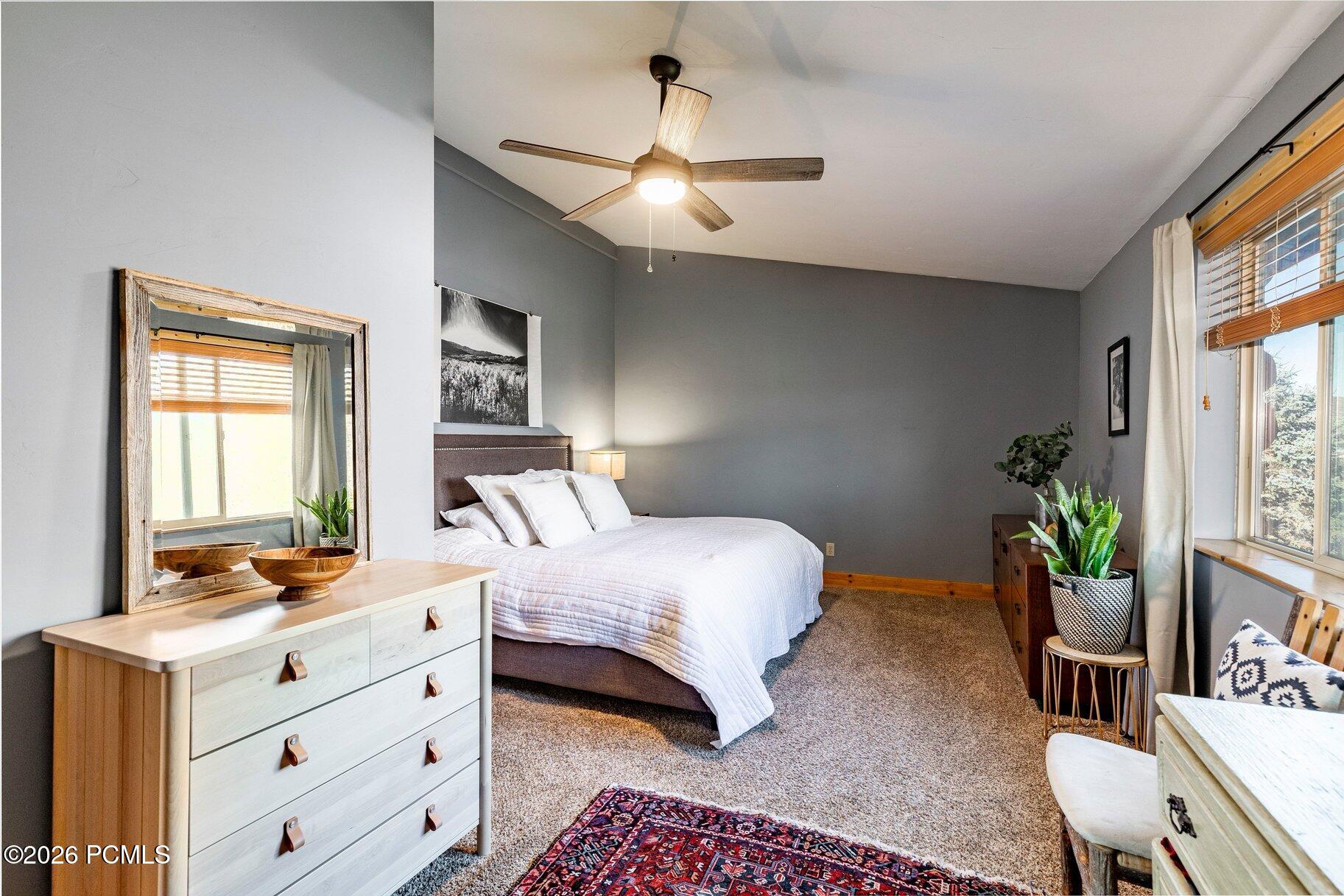 Furnished bedroom with a white bed, gray walls, ceiling fan, and large windows with wooden blinds.