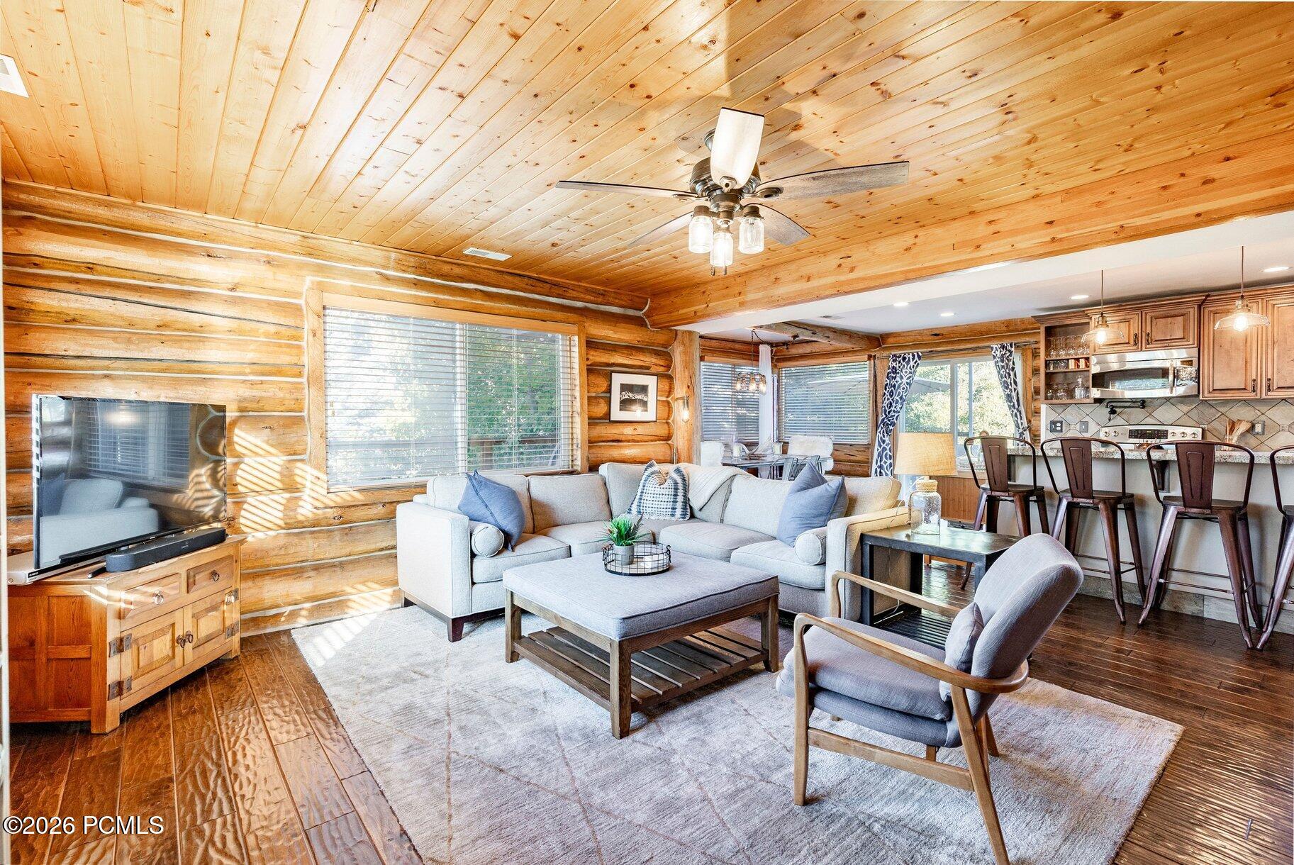 Furnished log-cabin living room with a pale sectional, wood ceiling, and open kitchen.