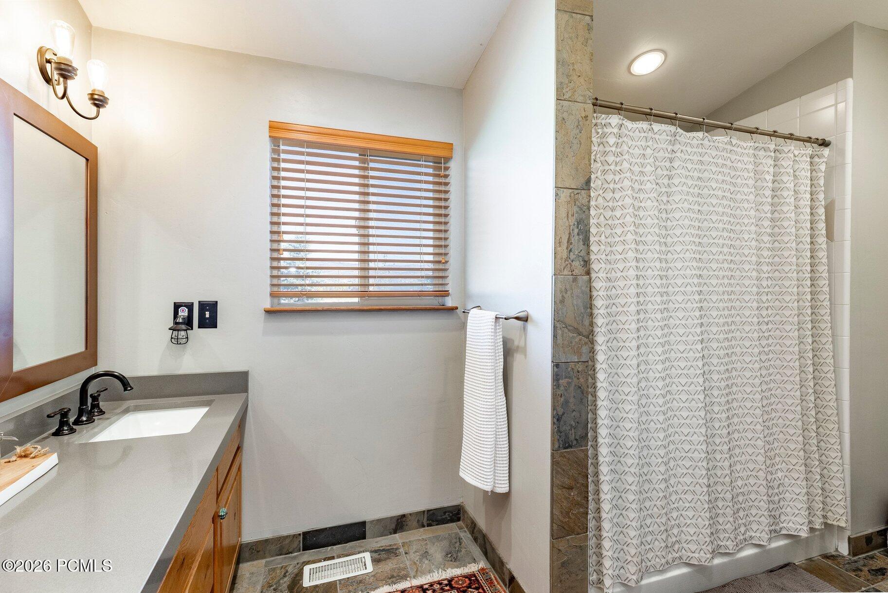 Bathroom with gray vanity, wood-framed mirror, window blinds, and stone-tiled shower surround.