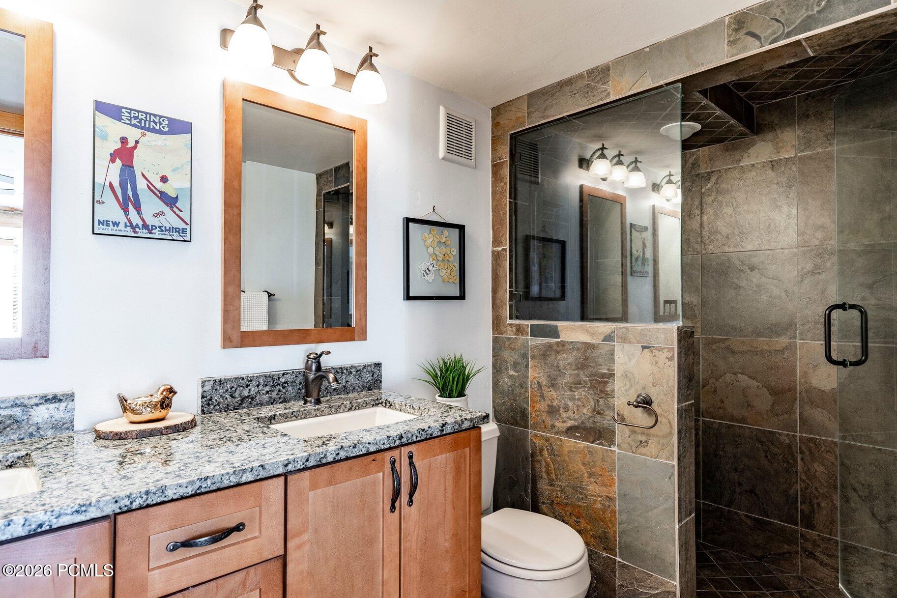 Bathroom with a granite vanity and wood cabinetry beside a glass-enclosed, stone-tiled shower.