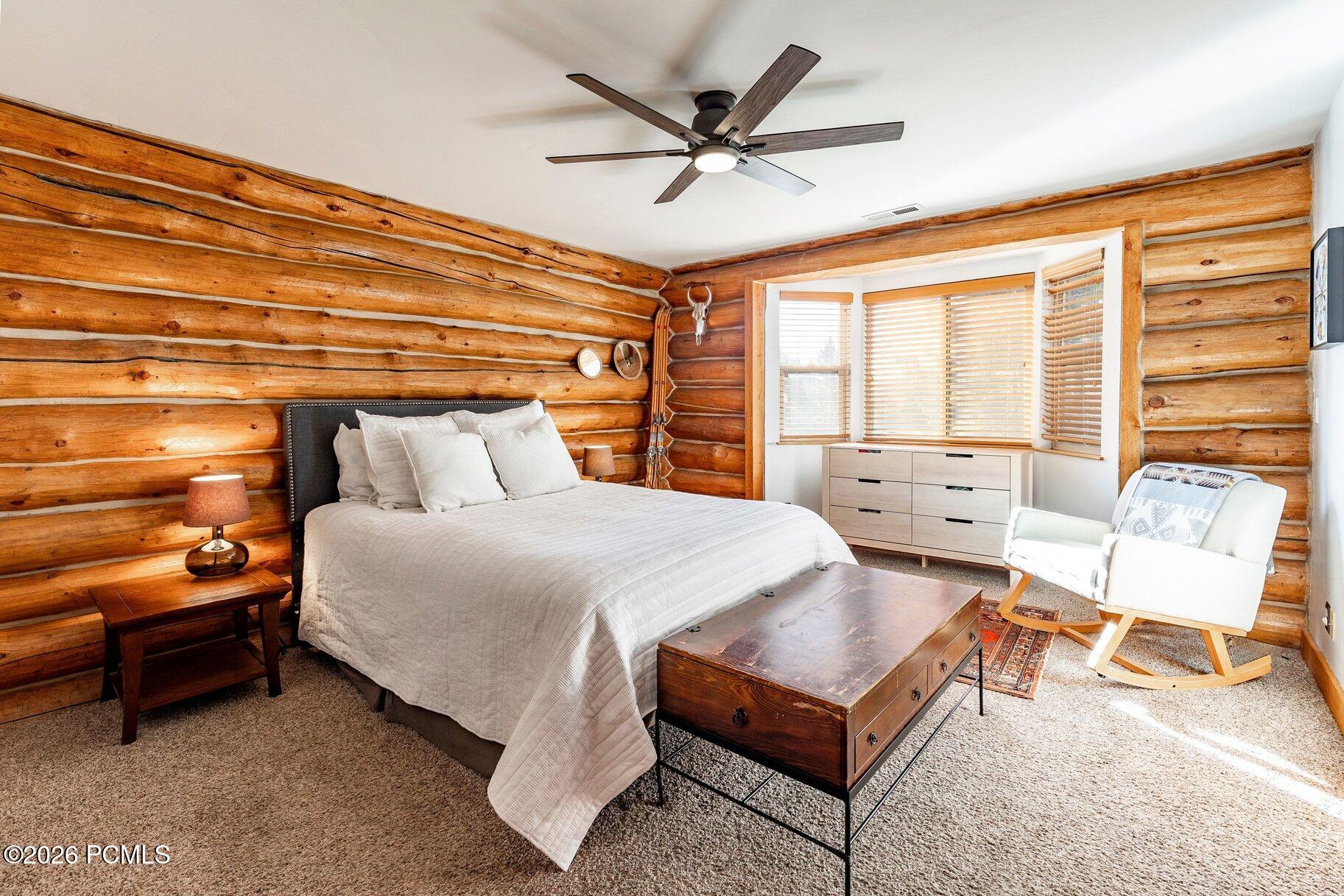 Bedroom with log walls, a white bed, bay window, and white rocking chair.