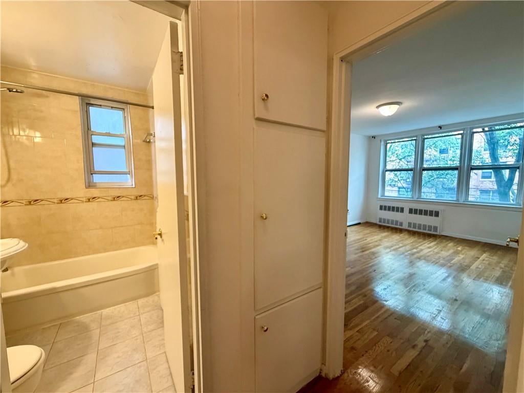 Vacant apartment interior with a tiled bathroom beside a bright hardwood-floored room with large windows.