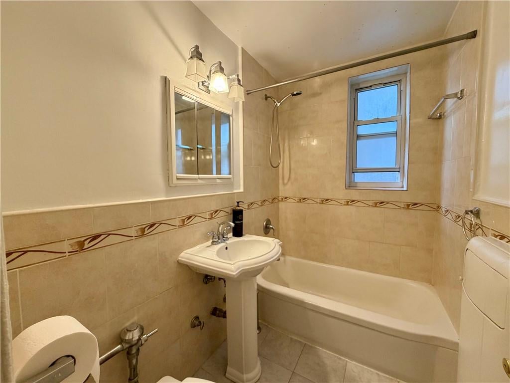 Bathroom with beige tiled walls, a bathtub and handheld shower, and a pedestal sink beneath a mirrored cabinet.