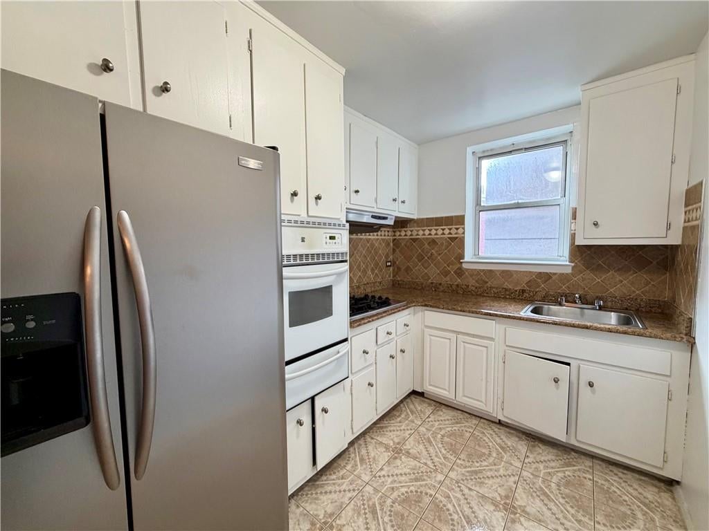 Kitchen with white cabinets, stainless refrigerator, and sink beneath a window; brown patterned tile covers the backsplash and floor.