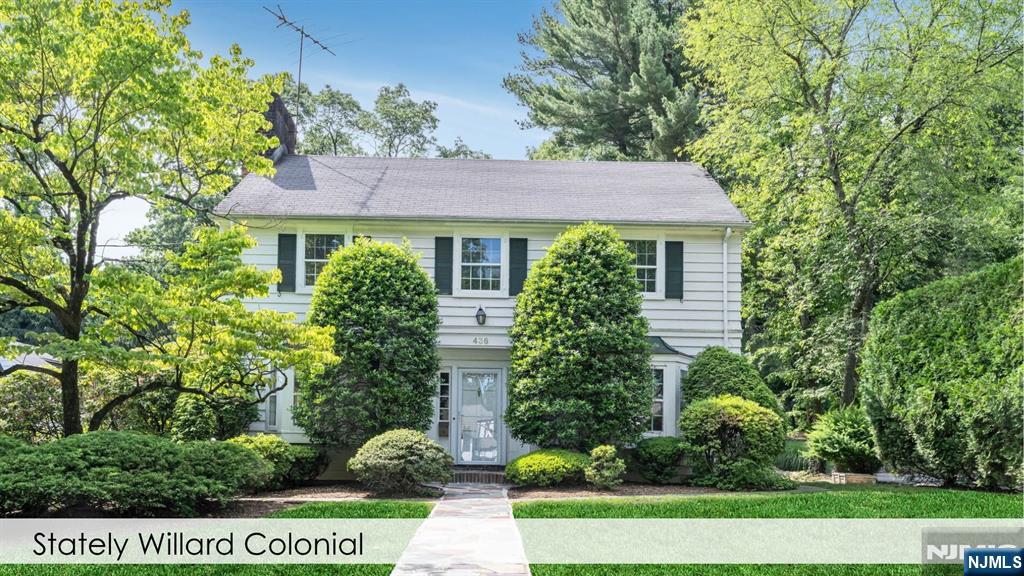 White two-story Colonial home with dark green shutters, mature trees, and a walkway to the front entrance.