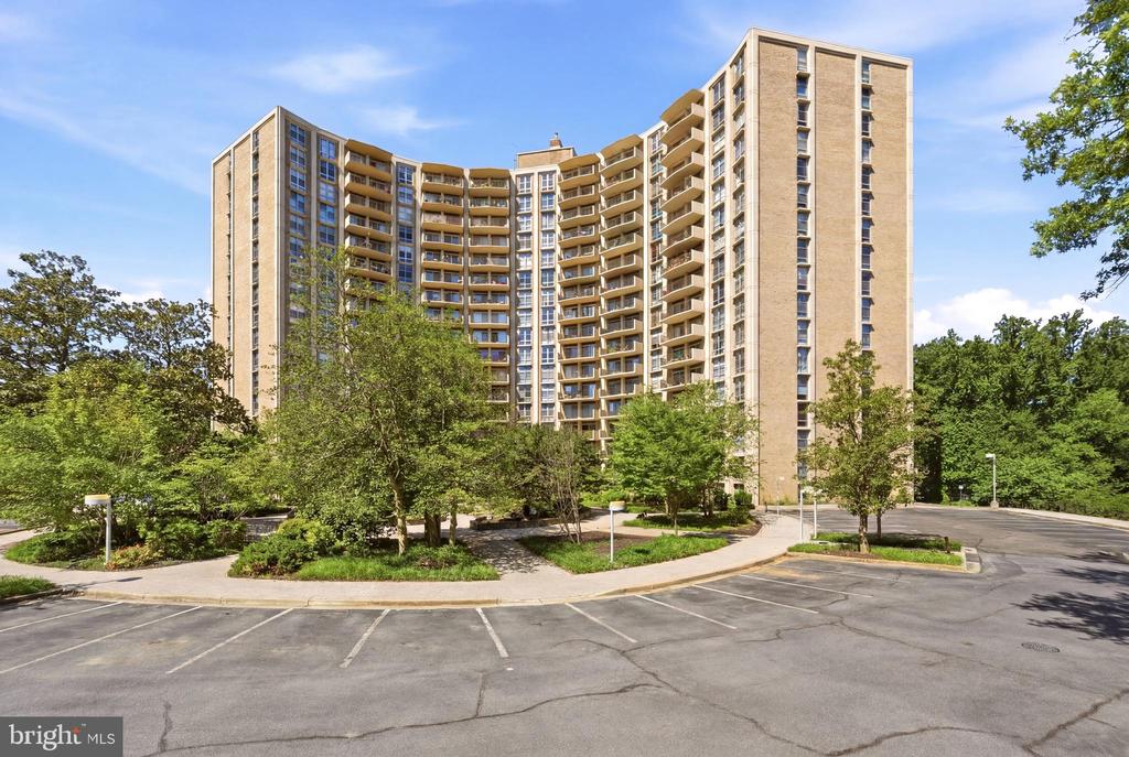 Curved beige-brick condominium building with balconies, trees, and a parking area.
