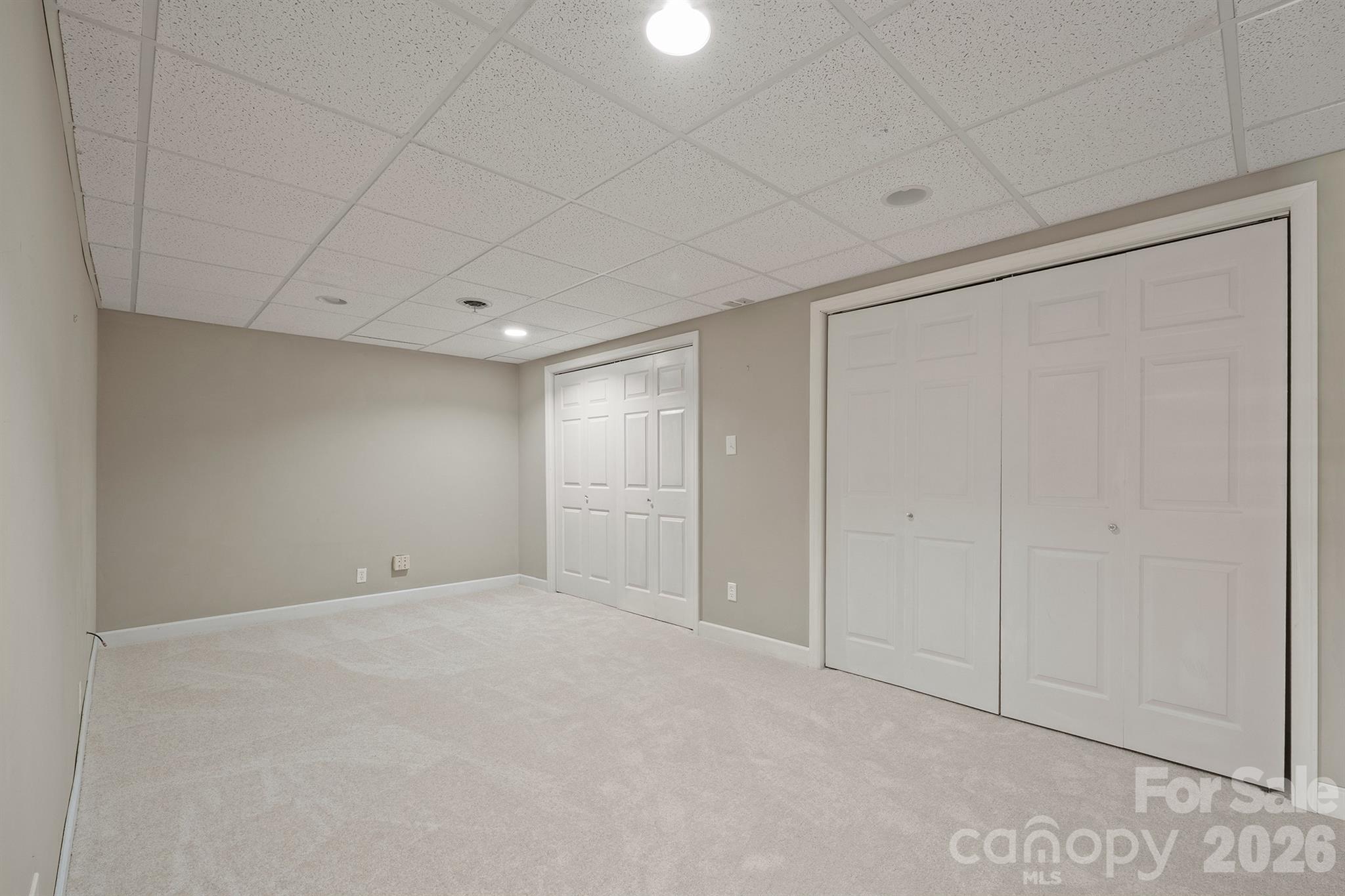 Empty carpeted room with beige walls, a tiled drop ceiling, and two sets of white closet doors.