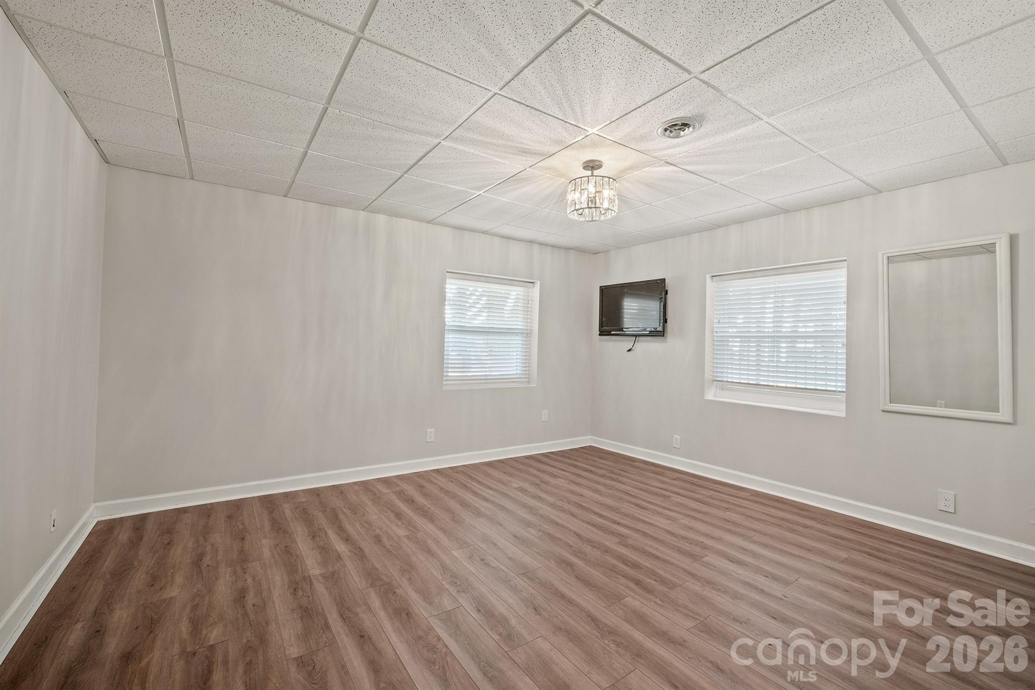 Empty room with wood-look flooring, two windows, a wall-mounted television, and a large mirror.