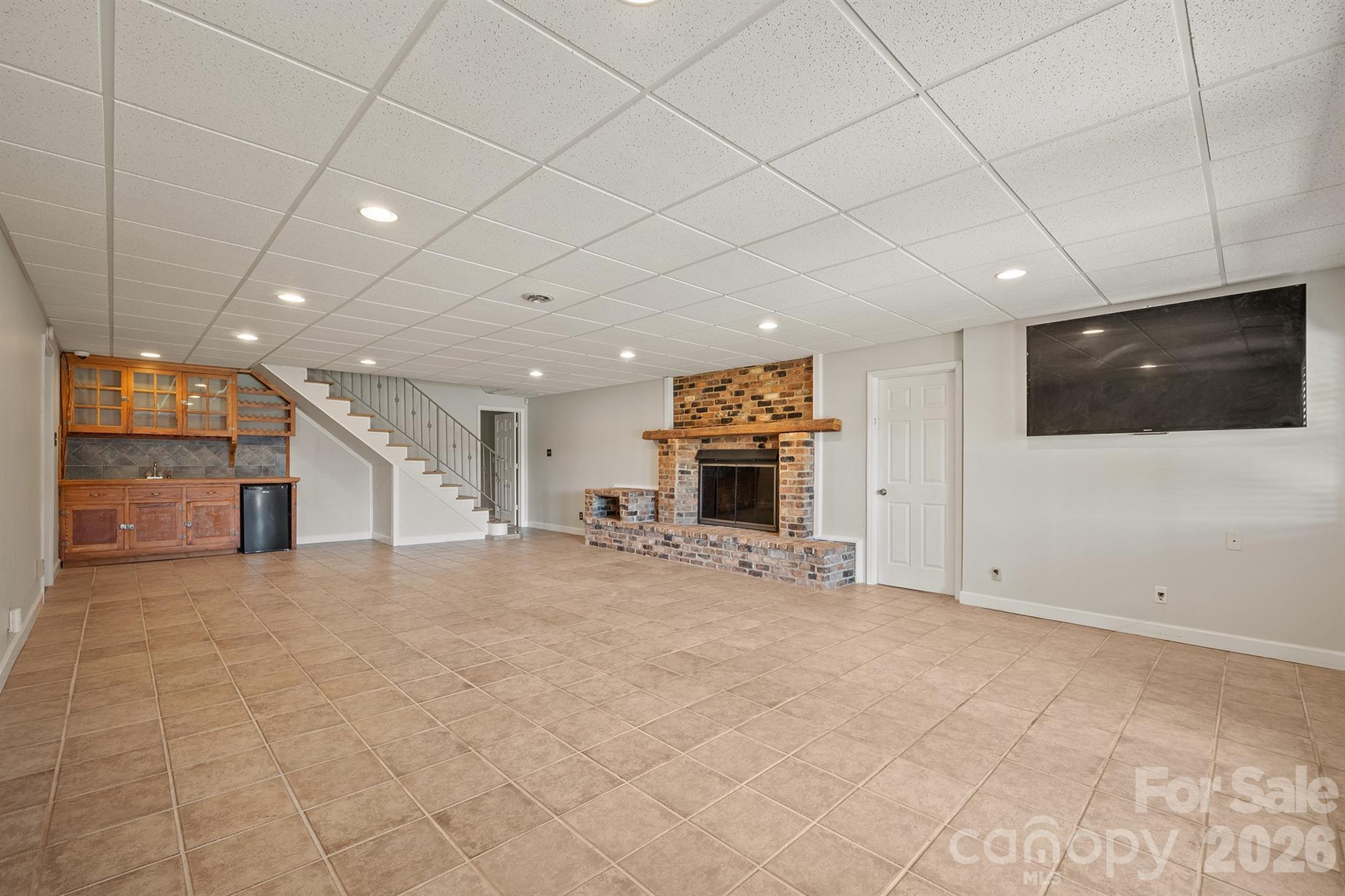 Finished basement recreation room with a brick fireplace, wooden wet bar, staircase, and wall-mounted TV.