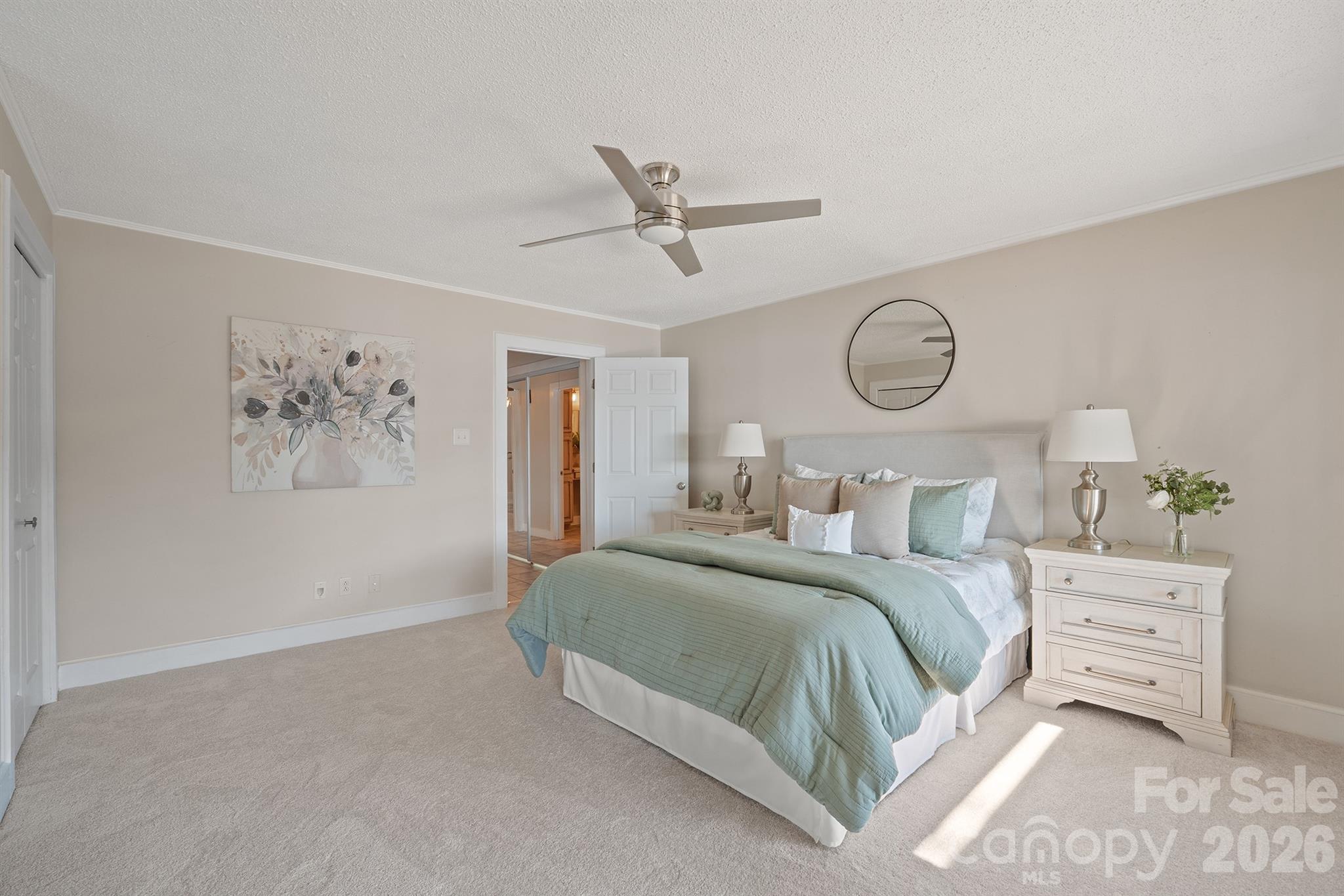 Staged bedroom with a king bed, pale sage bedding, ceiling fan, and round mirror.
