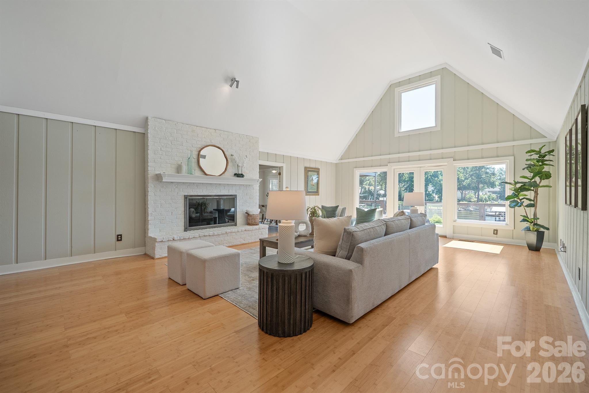 Furnished living room with vaulted ceiling, white brick fireplace, hardwood floors, and large windows facing a deck.