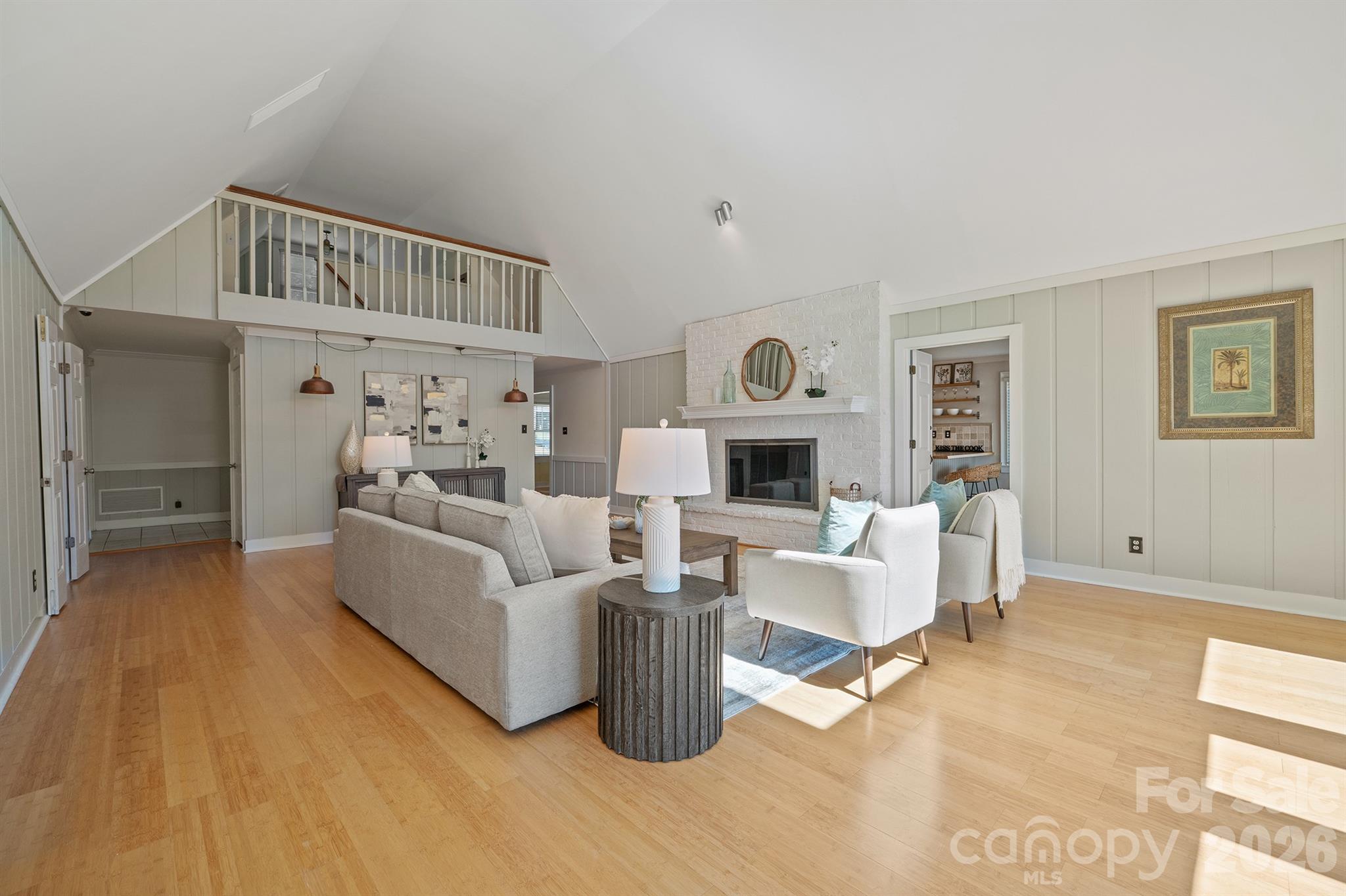 Furnished living room with vaulted ceiling, loft railing, and white brick fireplace.