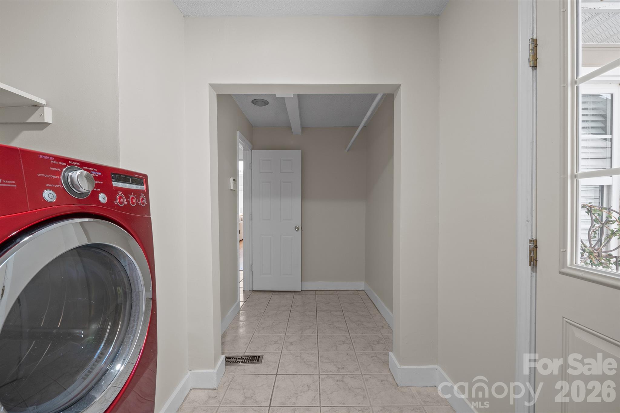 Laundry room with a red front-loading washer, tiled floor, and adjoining storage nook.