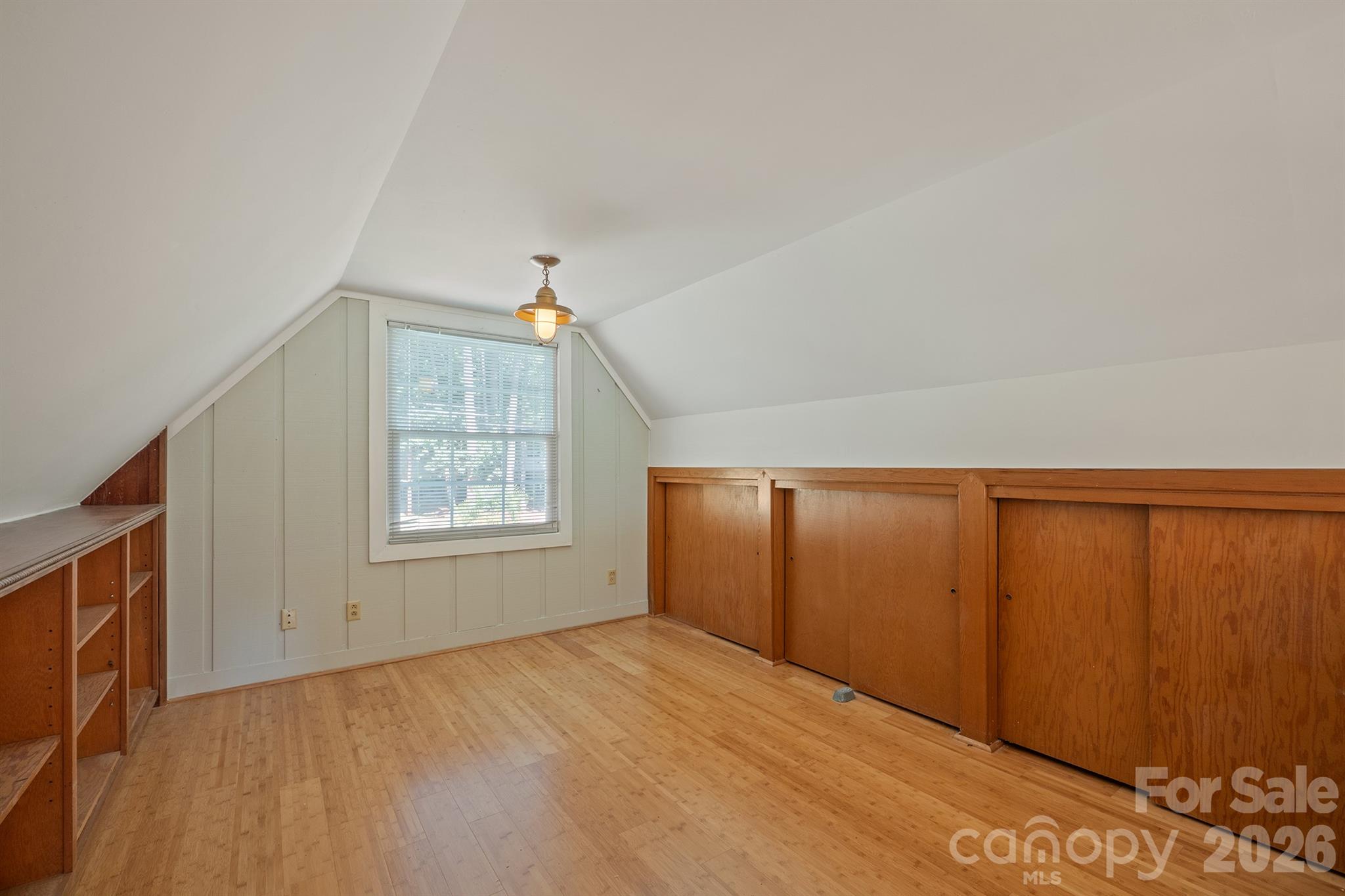 Empty attic bedroom with sloped ceilings, built-in wood cabinets, and a window with blinds.