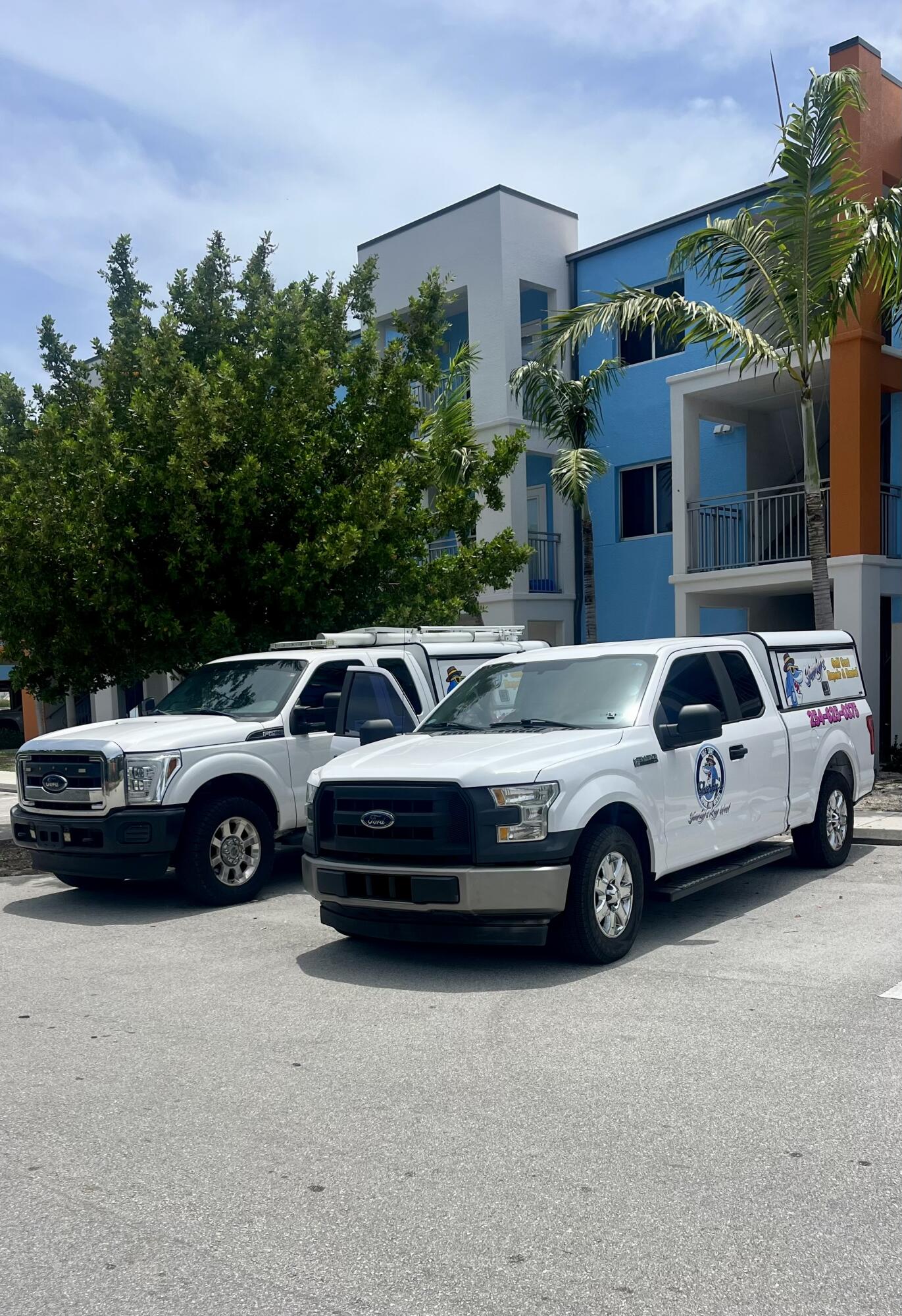 Two white service trucks parked outside a modern blue commercial building with palm trees.
