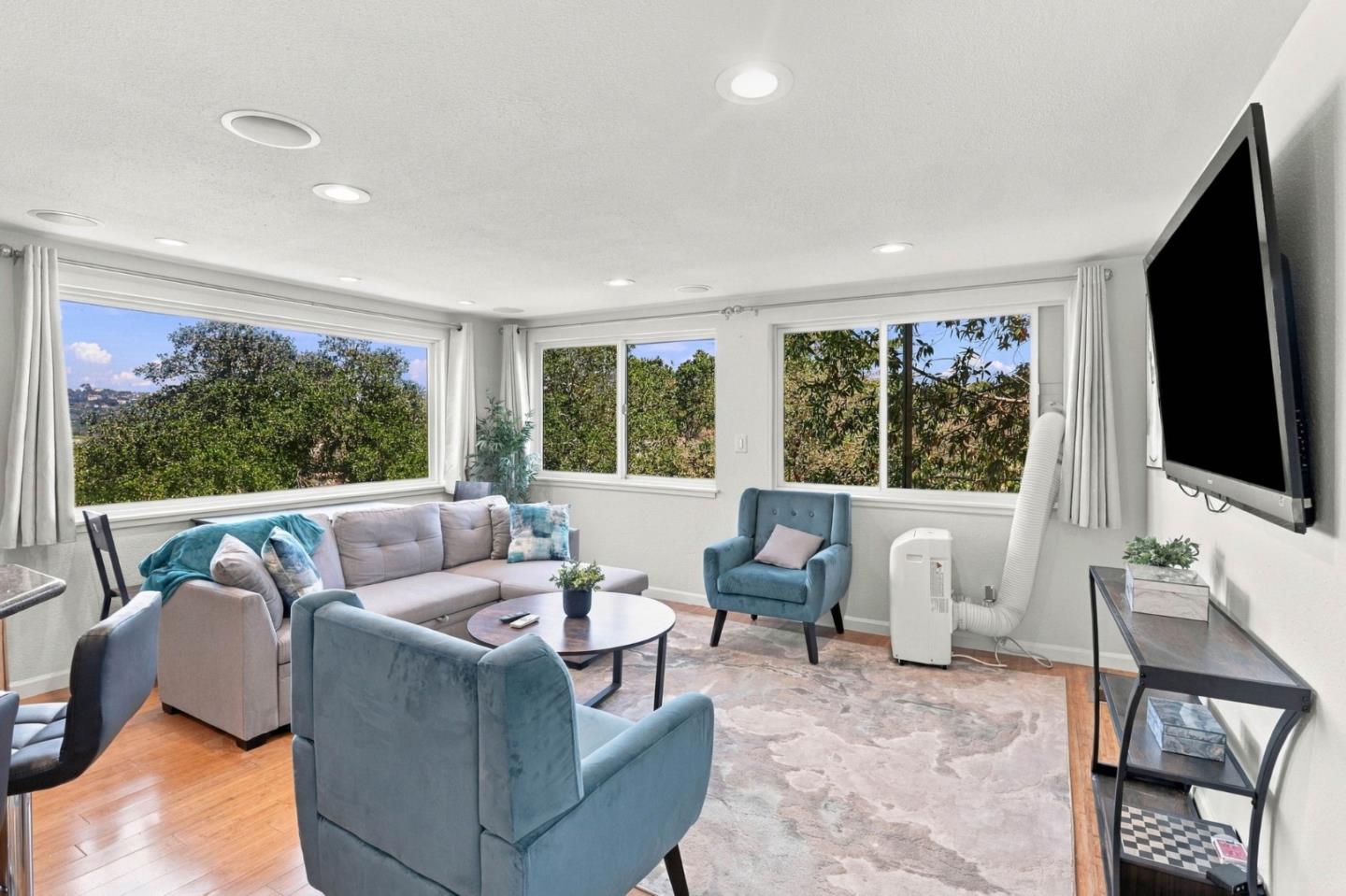 Furnished living room with wide windows overlooking leafy trees and teal accent chairs.