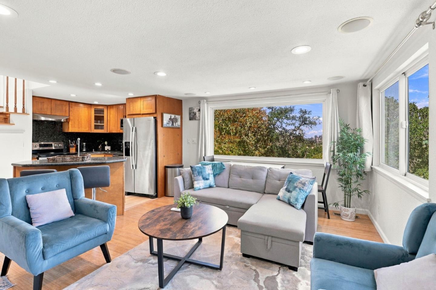 Open-plan living room and kitchen with wood cabinetry, a gray sectional, blue armchairs, and large windows.