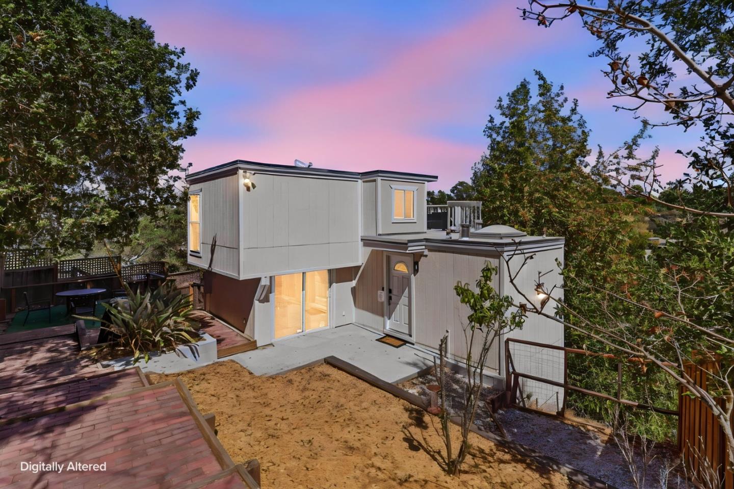 Modern white home with illuminated windows, a concrete patio, and a raised deck among trees at dusk.