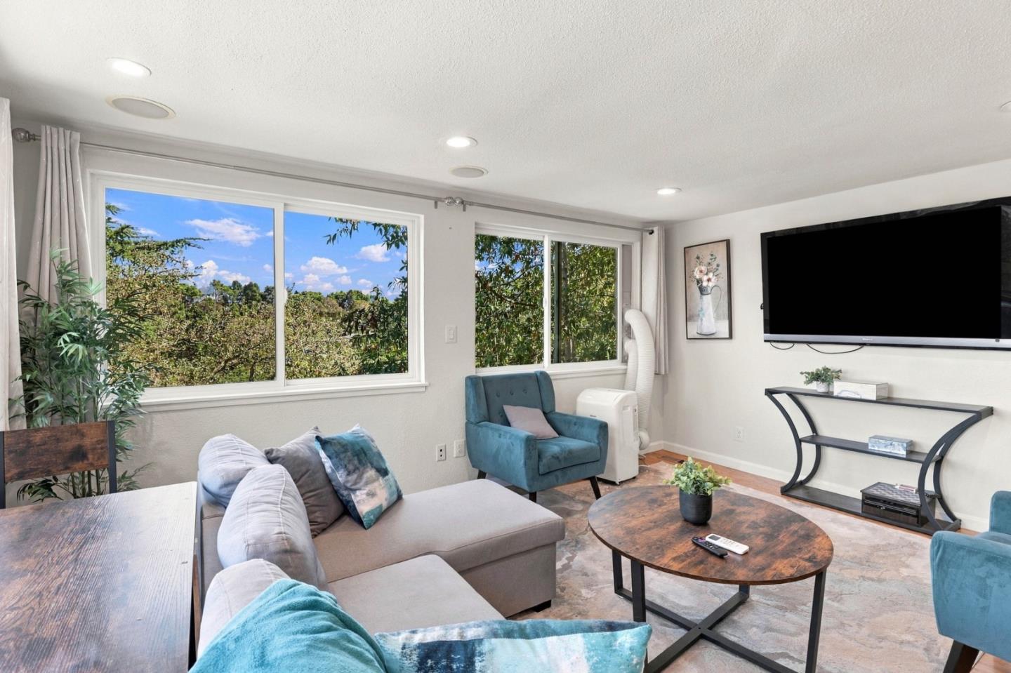Living room with a light sectional, teal chairs, round wood coffee table, and large windows overlooking trees.
