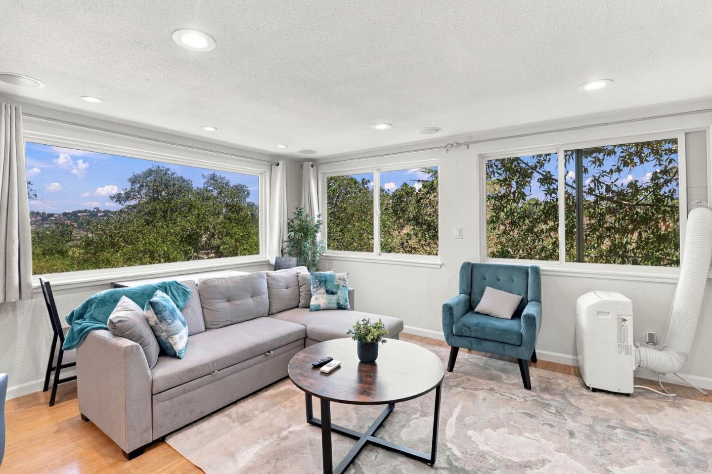 Living room with a gray sectional, teal armchair, and large windows overlooking trees and a hillside.