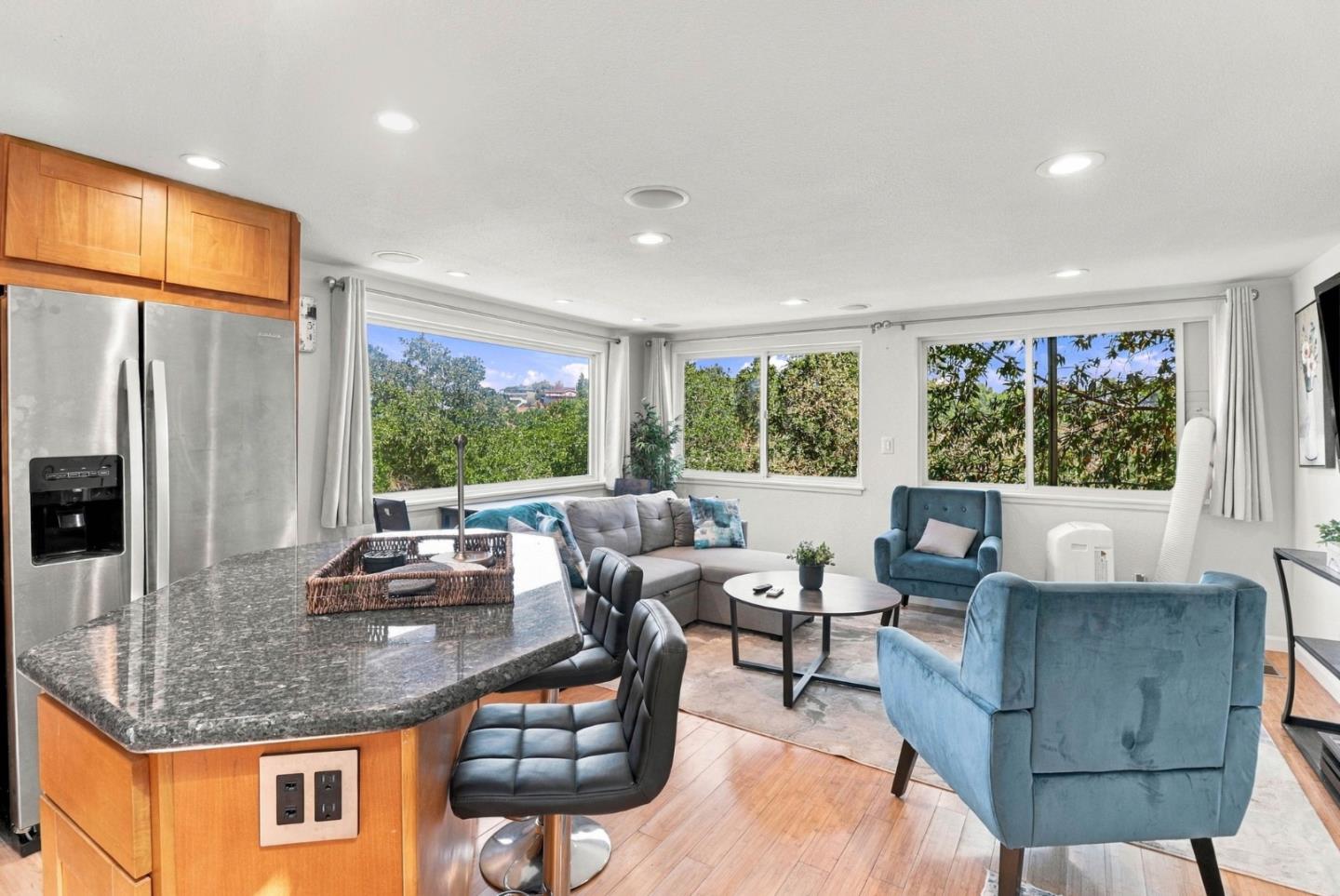 Open-plan kitchen and living room with a granite island, gray sectional, and blue armchairs by large windows.