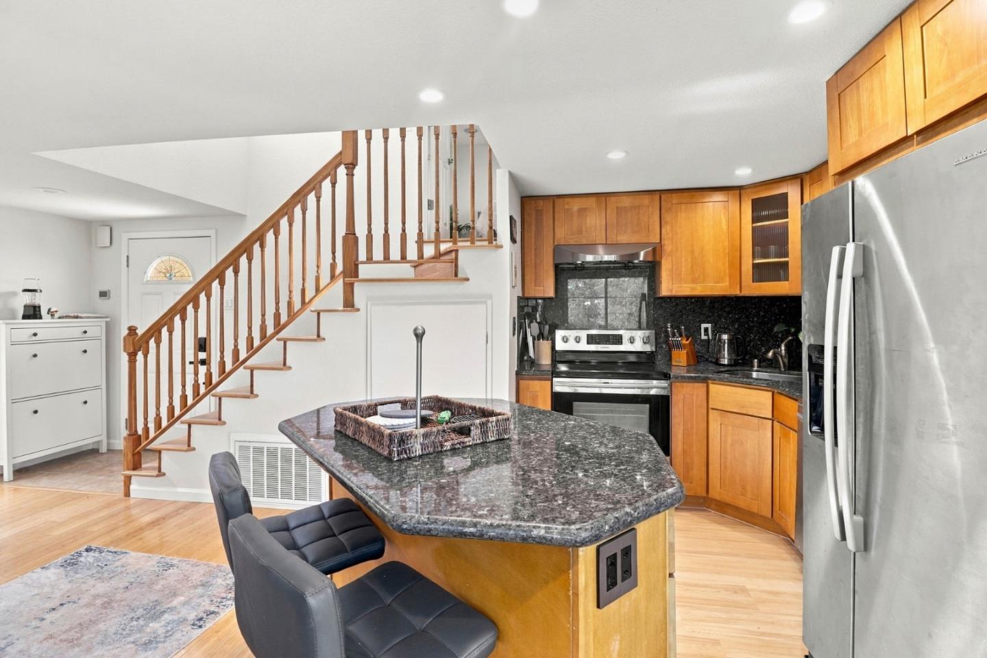 Kitchen with a granite island, warm wood cabinetry, stainless steel appliances, and an adjacent wooden staircase.