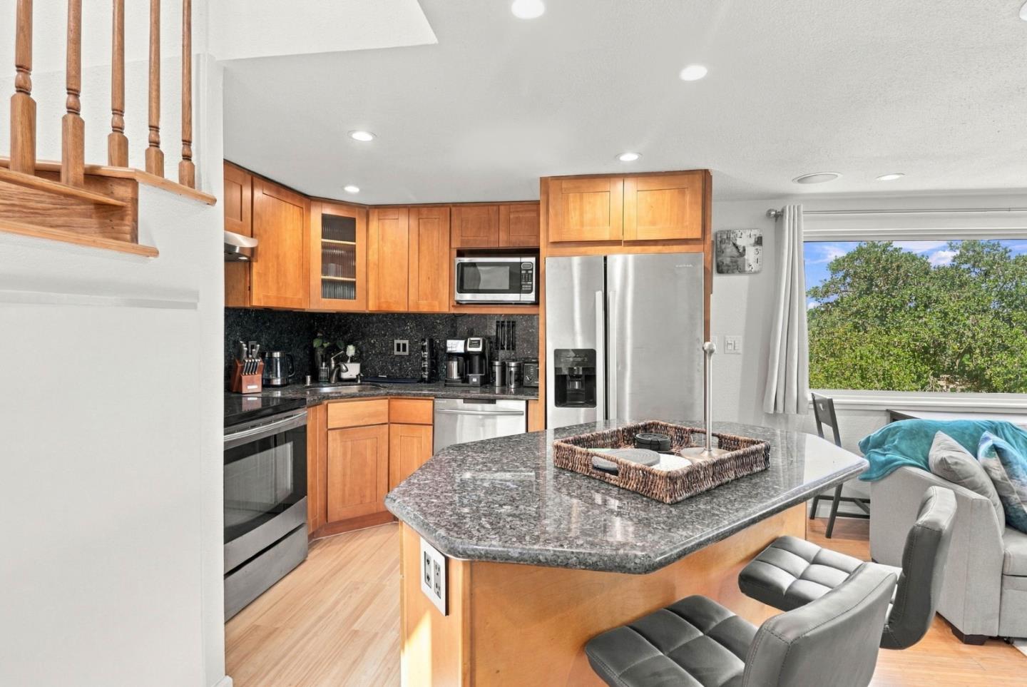 Kitchen with warm wood cabinets, granite island, stainless steel appliances, and a window overlooking trees.