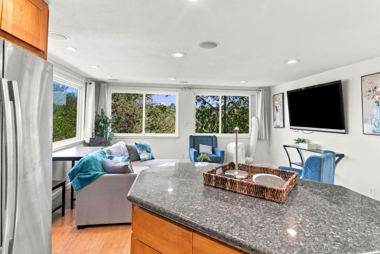 Kitchen and living area with a granite island, blue seating, and wide windows overlooking greenery.