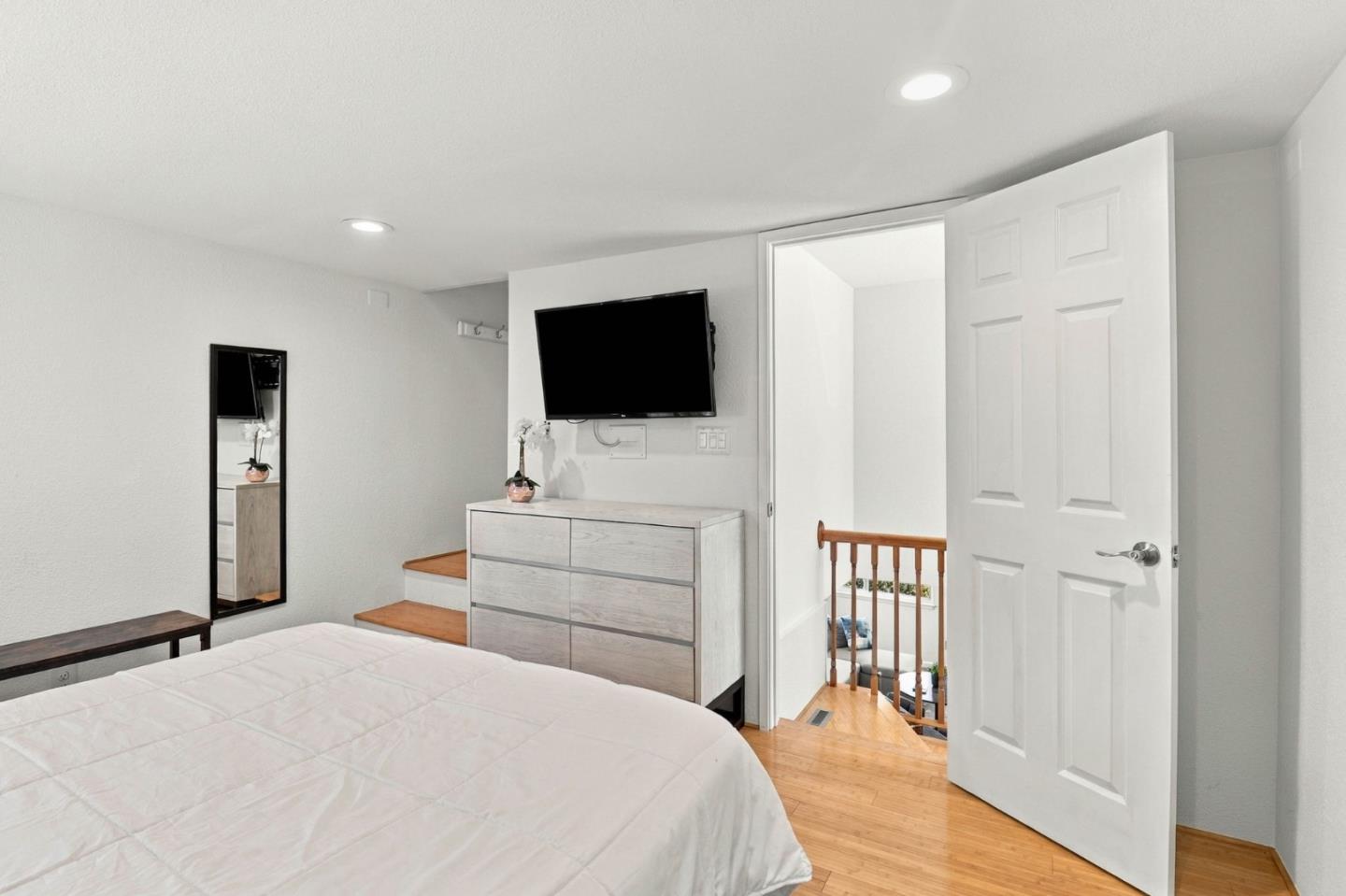 Bedroom with a white bed, wall-mounted television, light wood dresser and open door to the stair landing.