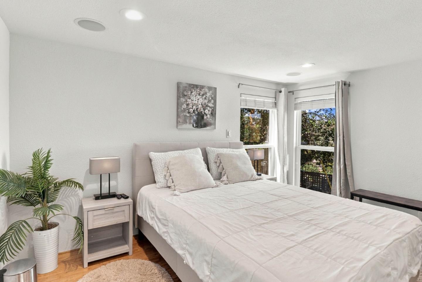 Bright bedroom with a large bed, potted palm, and two windows overlooking greenery.