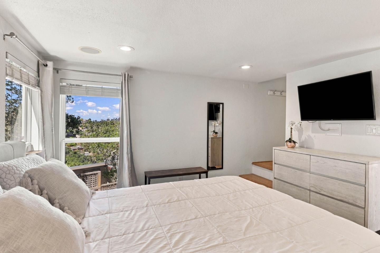 Bedroom with a large bed, wall-mounted TV, and windows overlooking leafy trees.
