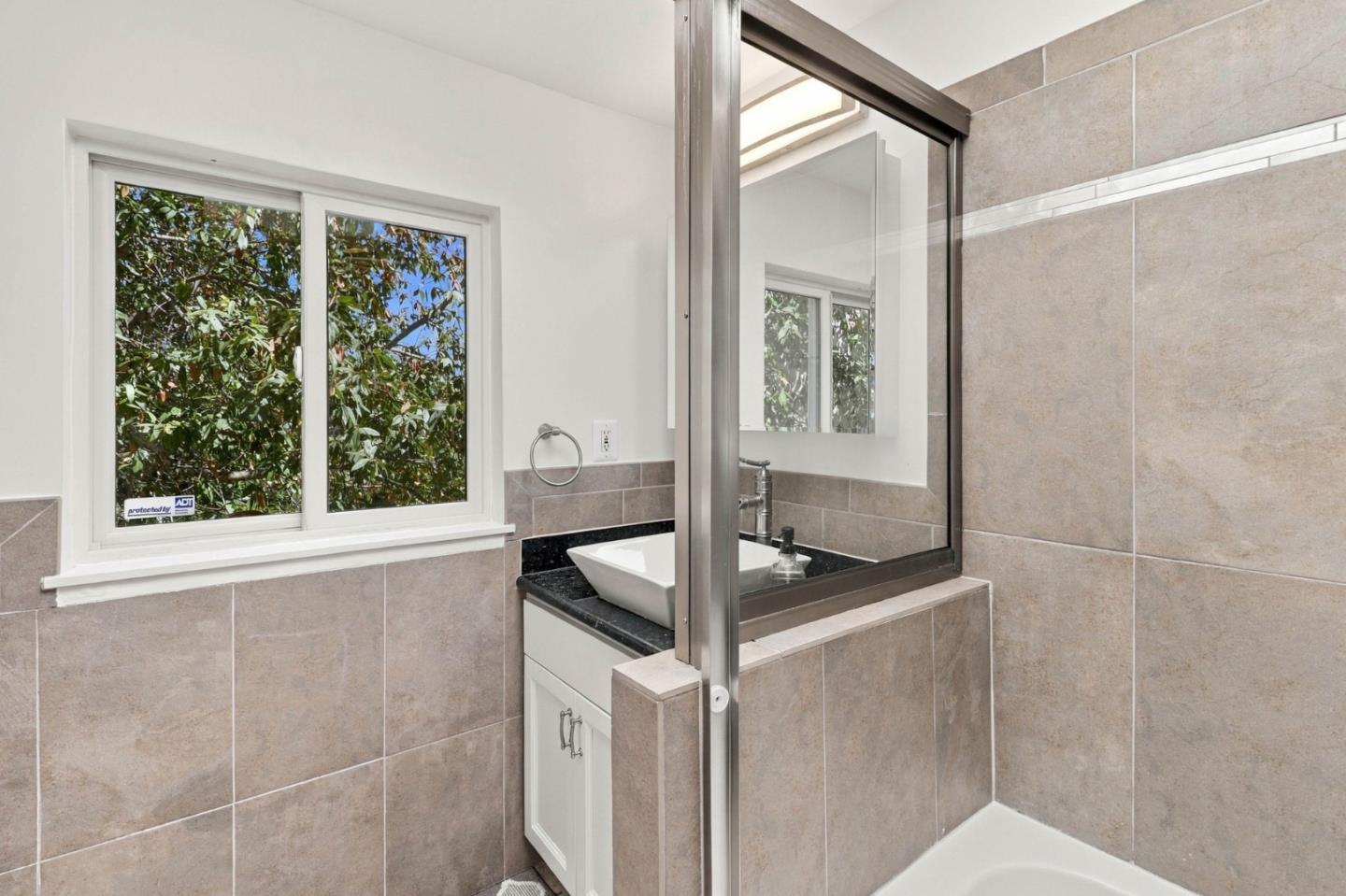 Bathroom with tiled shower enclosure, white vessel sink, and window overlooking leafy trees.