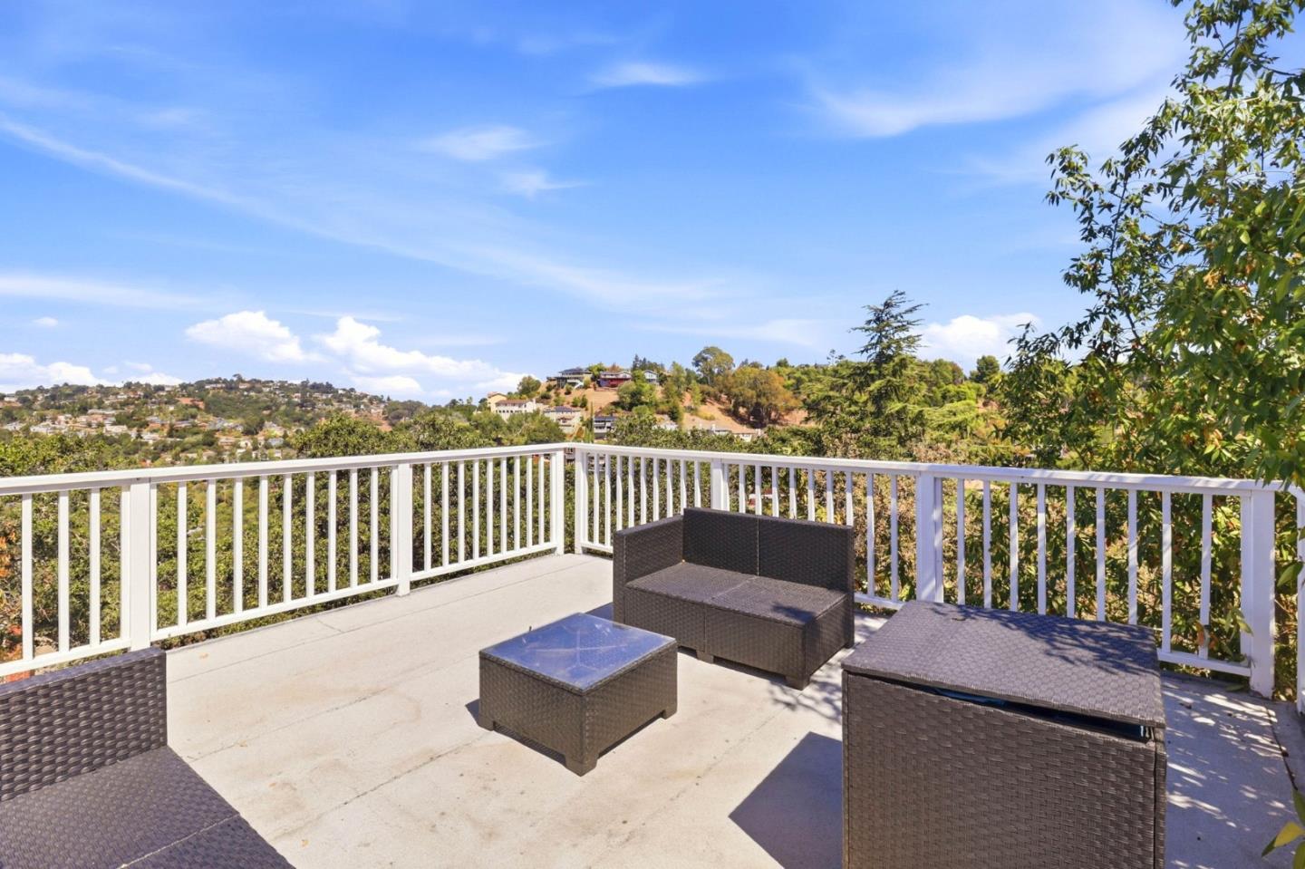 Furnished deck with dark wicker seating and white railing overlooking wooded hills and homes.