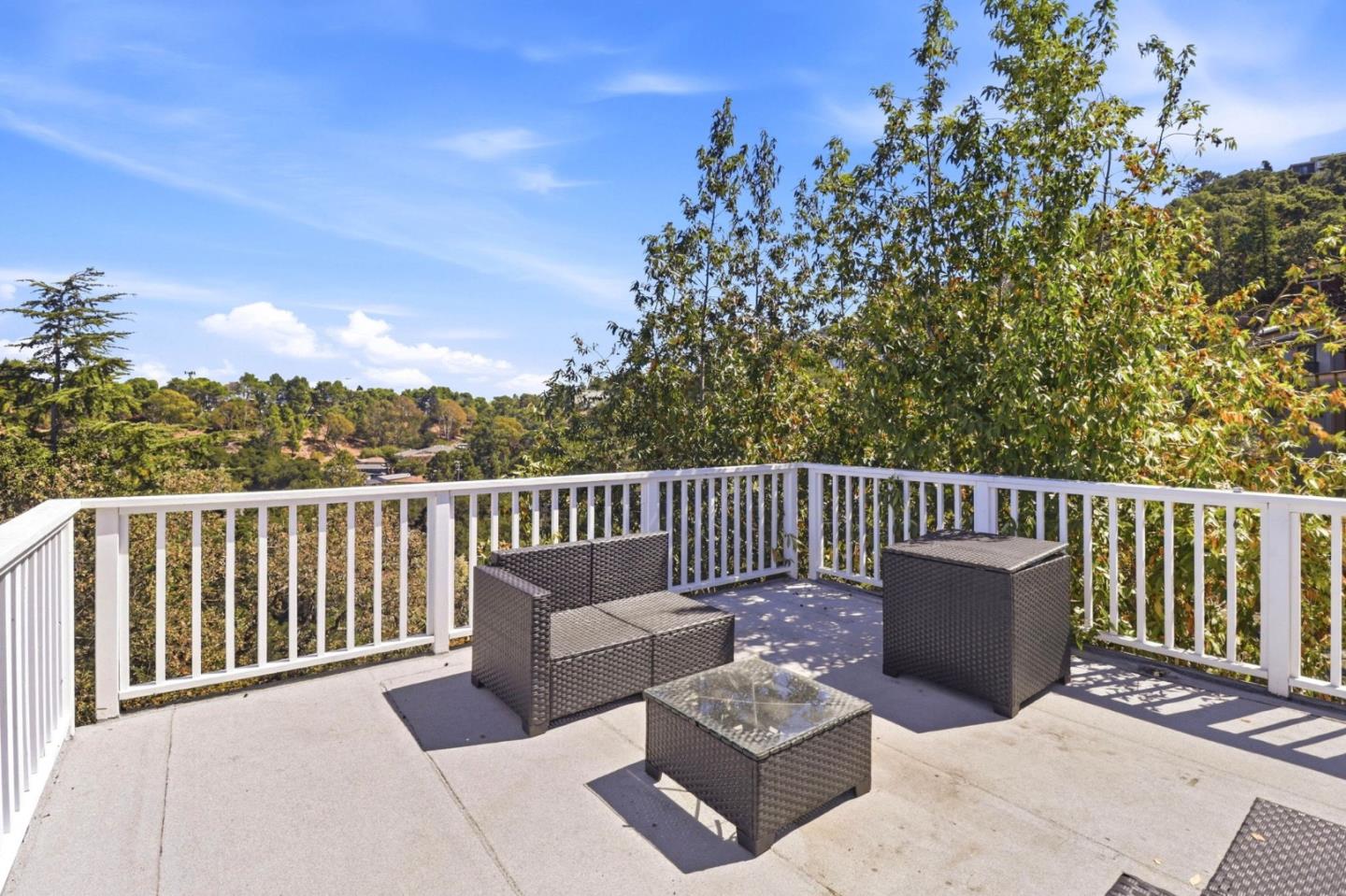 Furnished deck with white railings, woven patio seating, and a view of leafy hills.