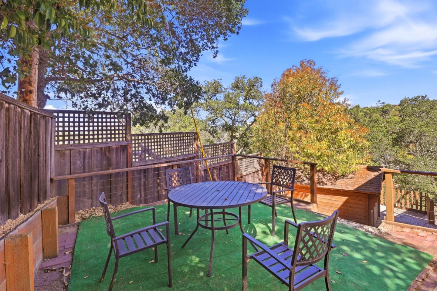 Outdoor deck with round table and chairs, green turf, and wooden lattice fencing beneath mature trees.