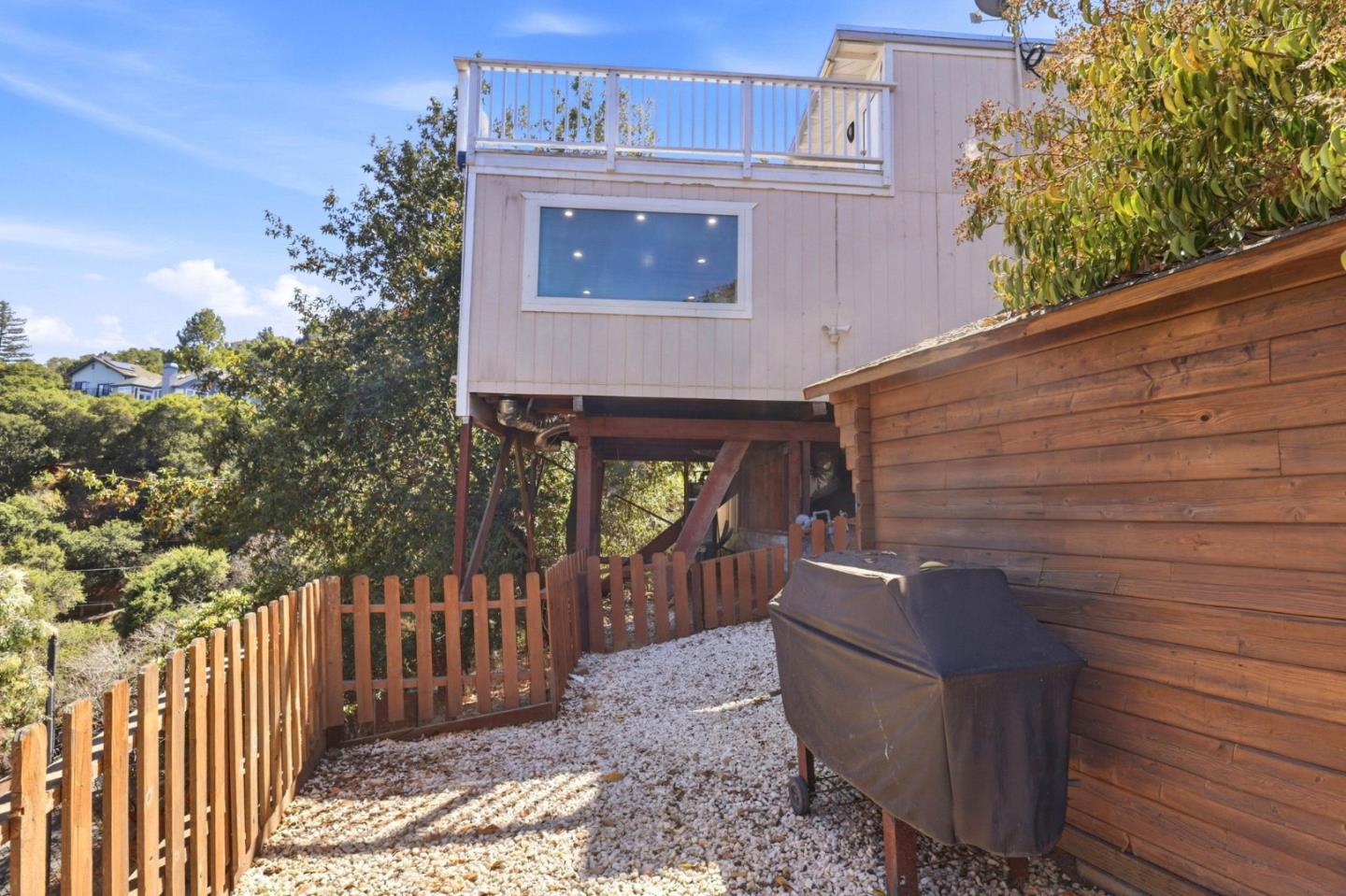 Raised house beside a gravel patio with a wooden fence, shed, and covered grill.