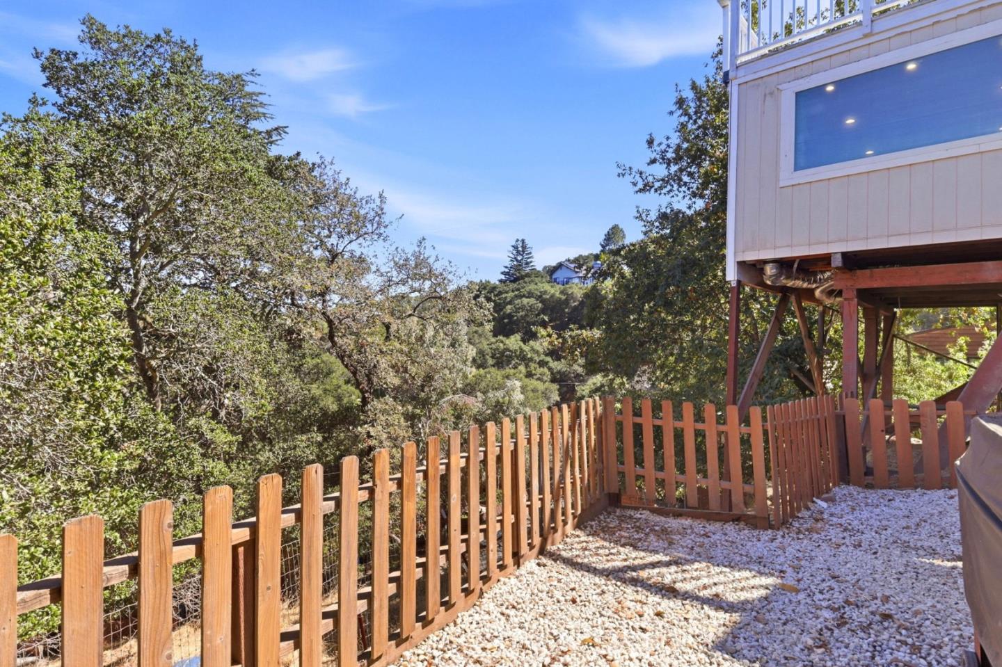 Side yard with a wooden fence, white gravel, and leafy trees beside a raised home.