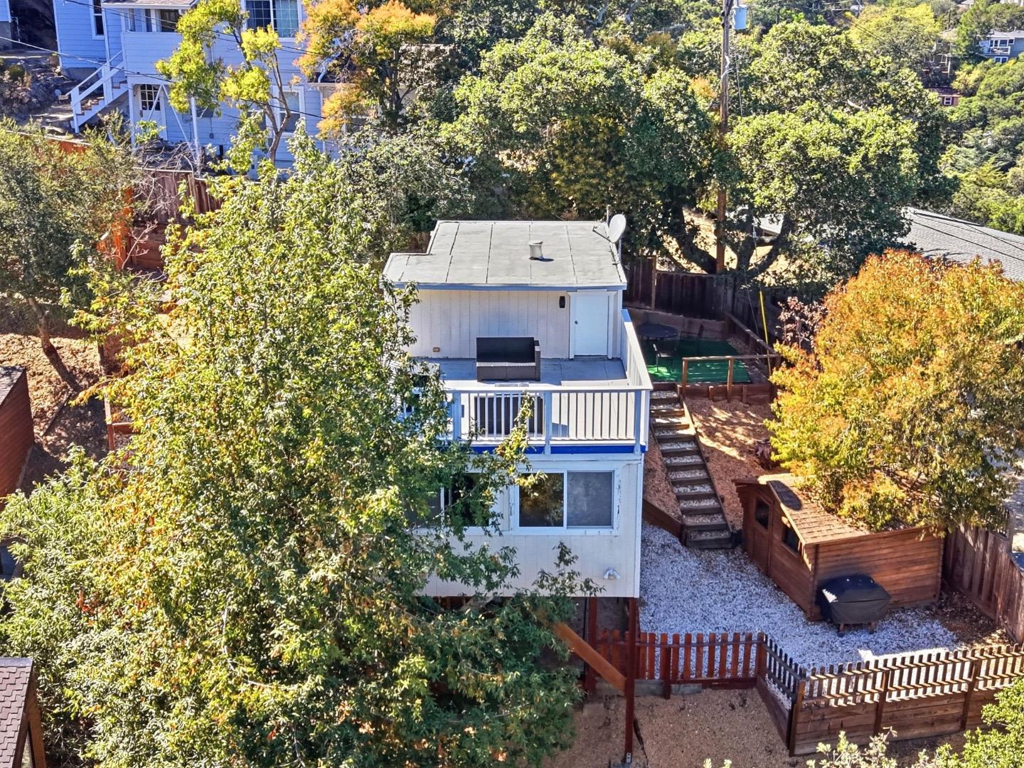 Raised backyard deck with a bench and stairs, surrounded by mature trees and a fenced gravel yard.